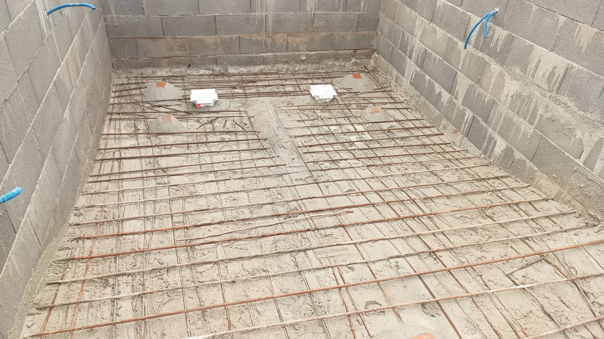 Canteiro de obras de uma piscina retangular: paredes de blocos de concreto cinza, tela de vergalhões no piso, canos brancos.