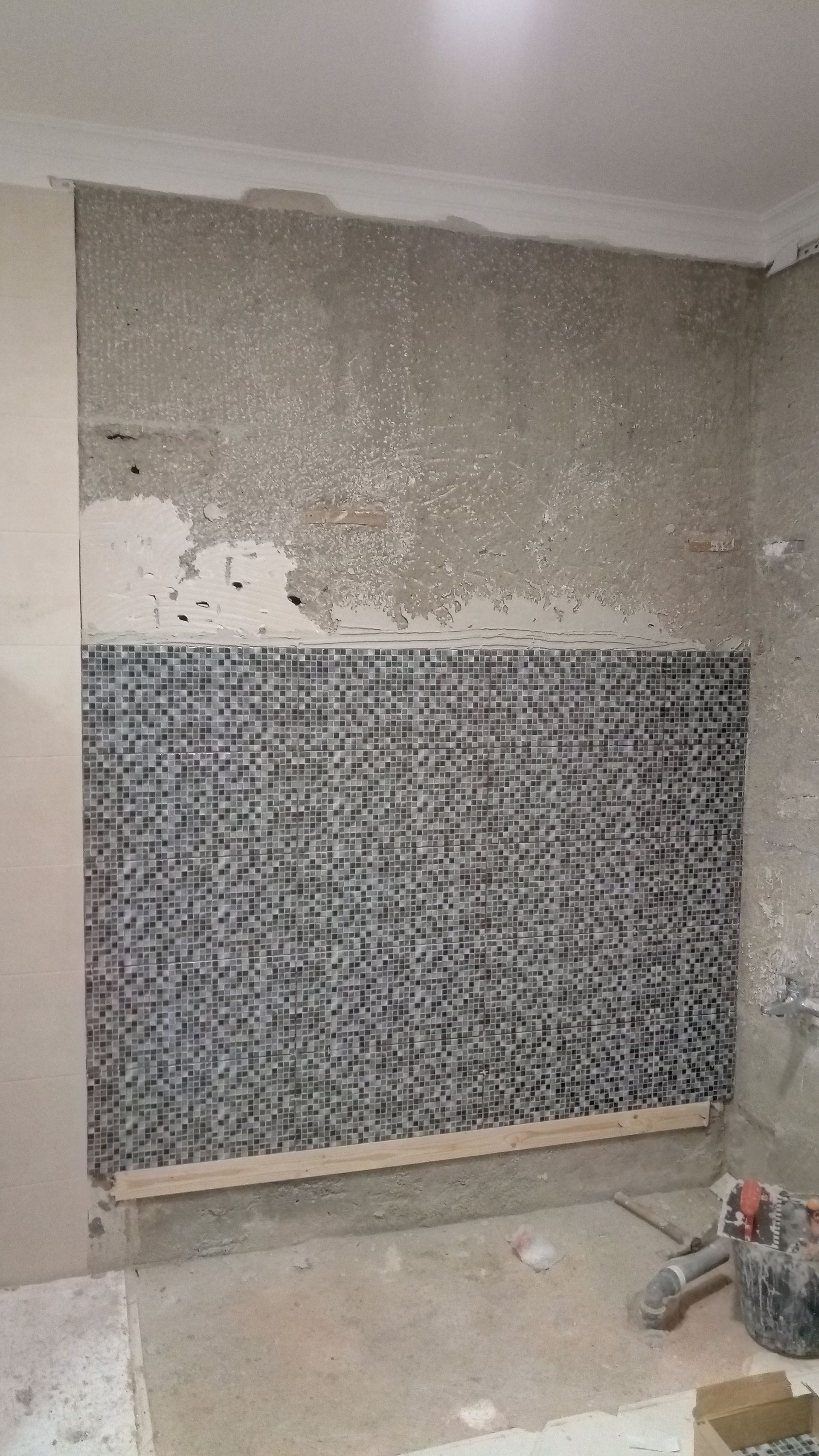 Parede do banheiro em construção, parcialmente revestida com azulejos. Azulejos de mosaico cinza, concreto aparente, suporte de madeira.