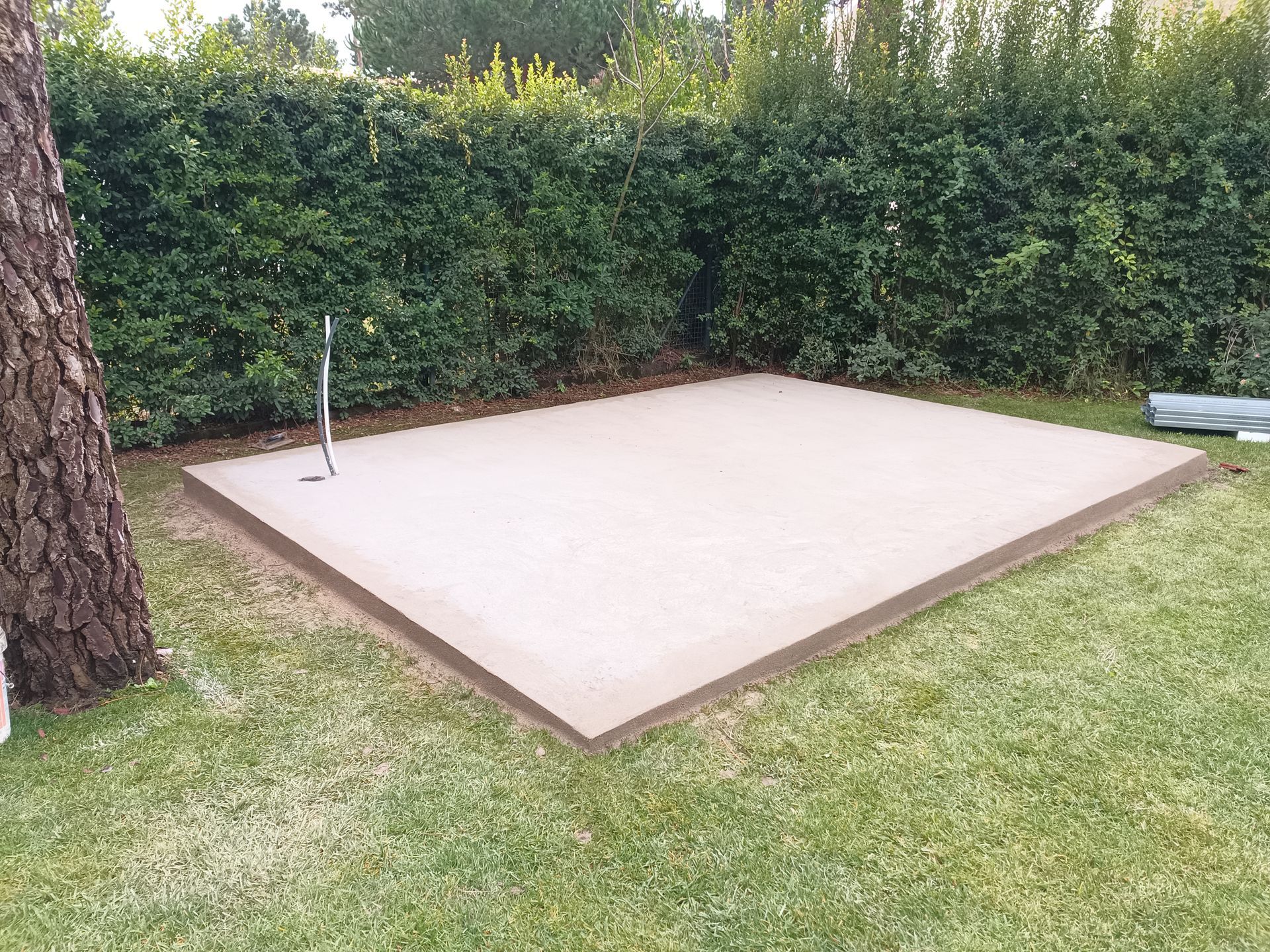 Uma plataforma quadrada de concreto com borda escura está localizada na grama, ao lado de uma árvore e uma cerca viva.