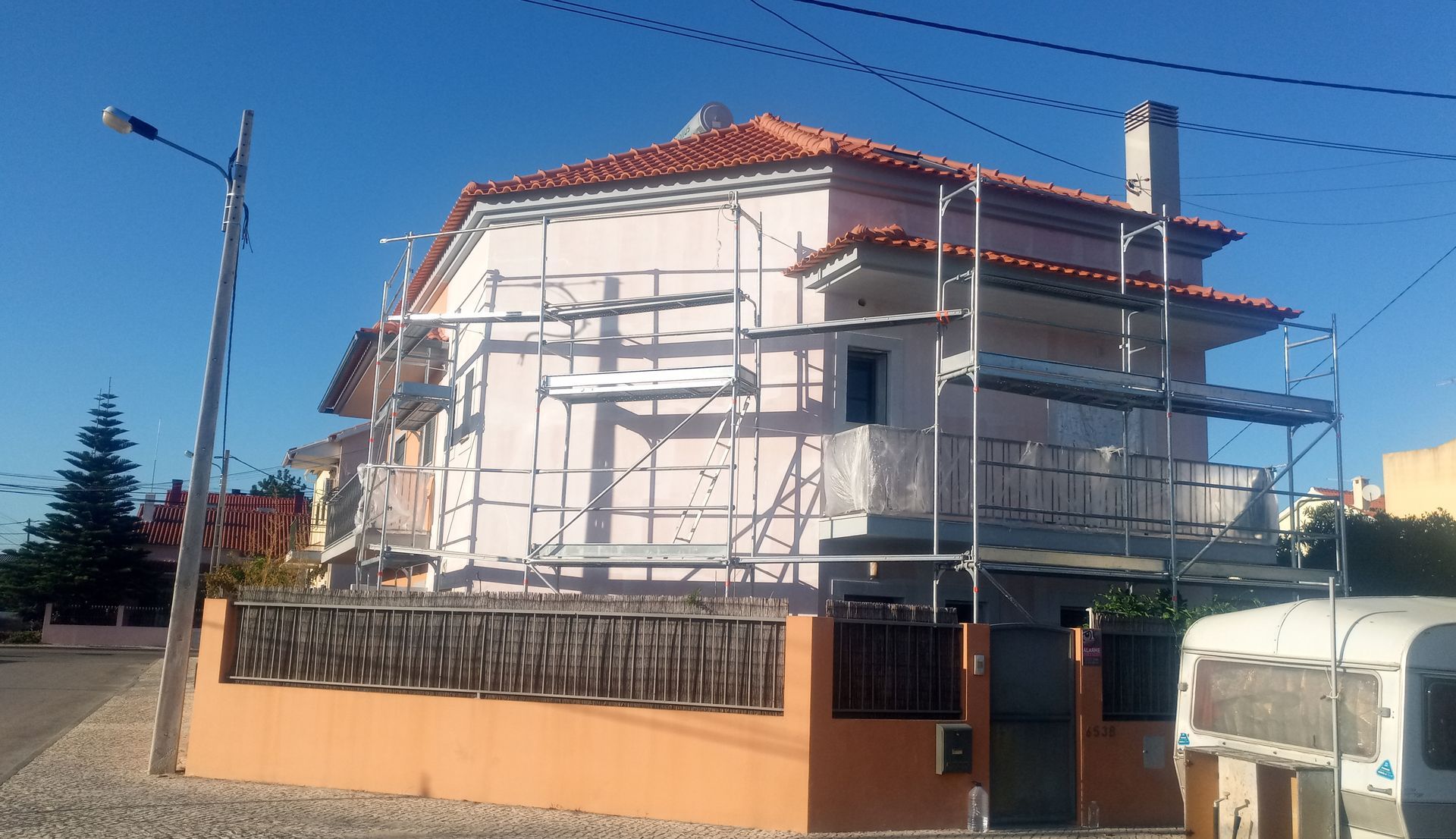 Casa em reforma com andaimes, paredes rosa e telhado de telha vermelha contra um céu azul.