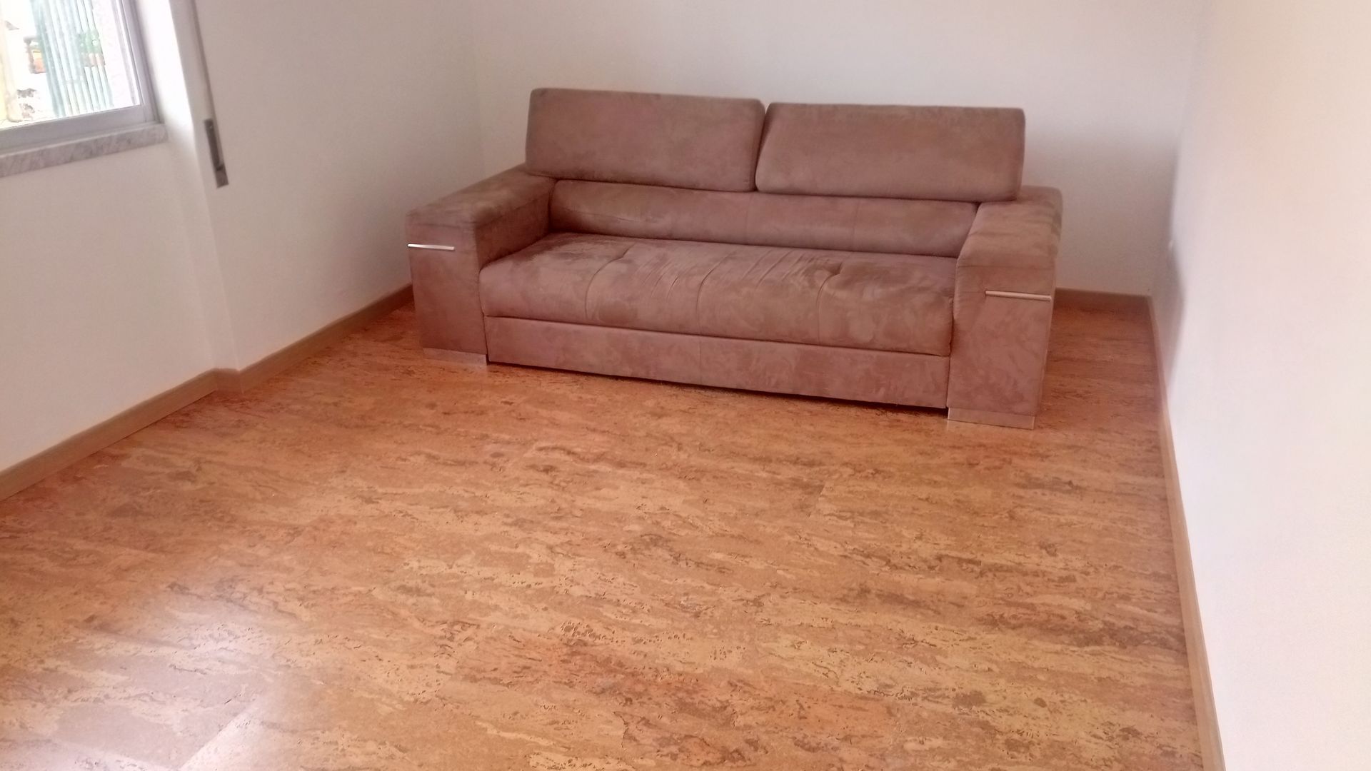 Sofá de dois lugares marrom em um quarto com mobília esparsa, piso que imita madeira e uma janela.