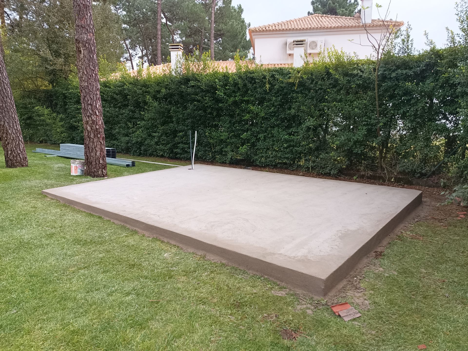 Plataforma de concreto recém-construída em um gramado, cercada por árvores e uma cerca viva.