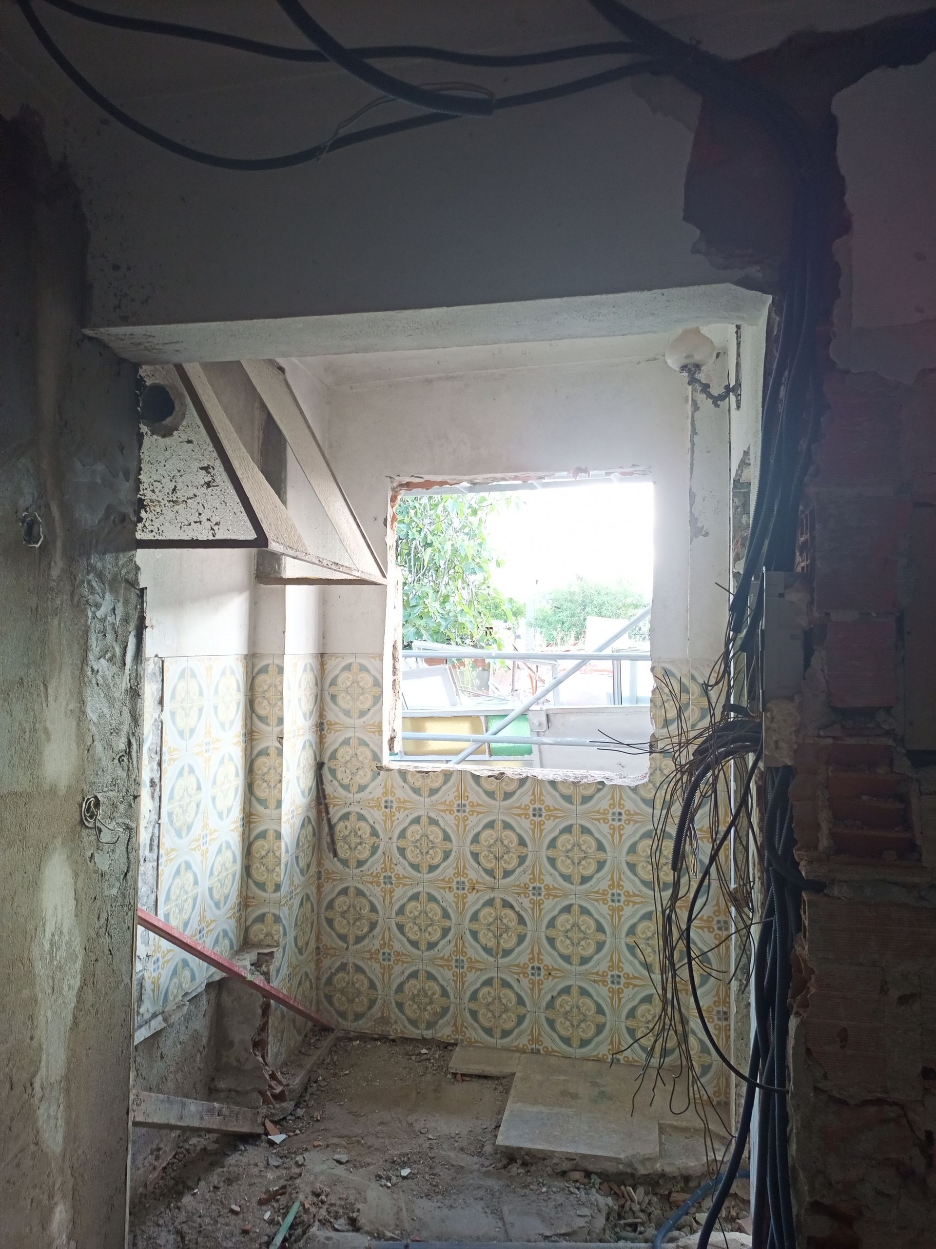 Demolição interna: cômodo com parede revestida de azulejos, fiação exposta e janela que dá para uma área externa arborizada.