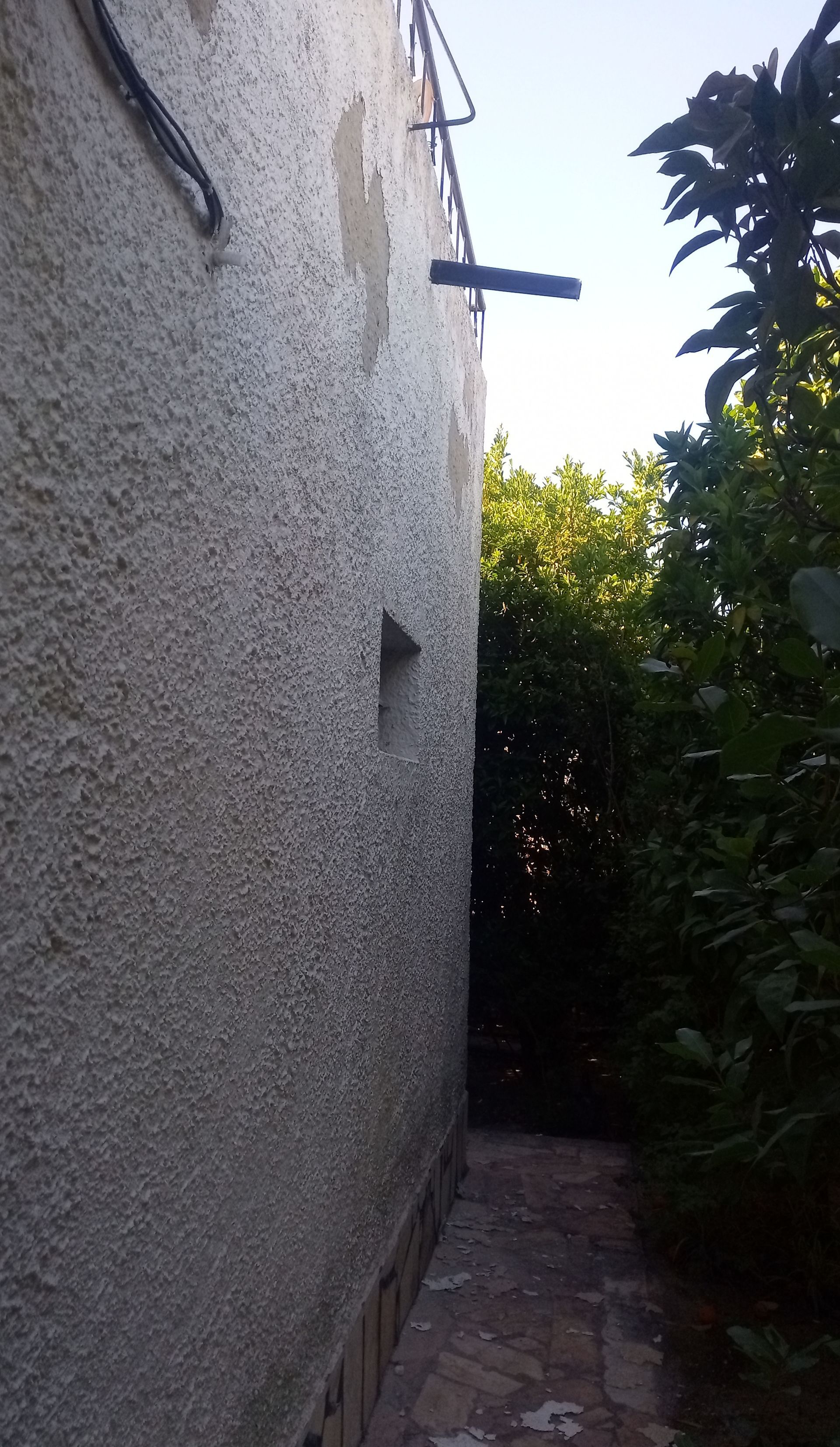 Parede externa texturizada de um edifício com uma pequena janela triangular e um caminho estreito que leva a uma área verde.