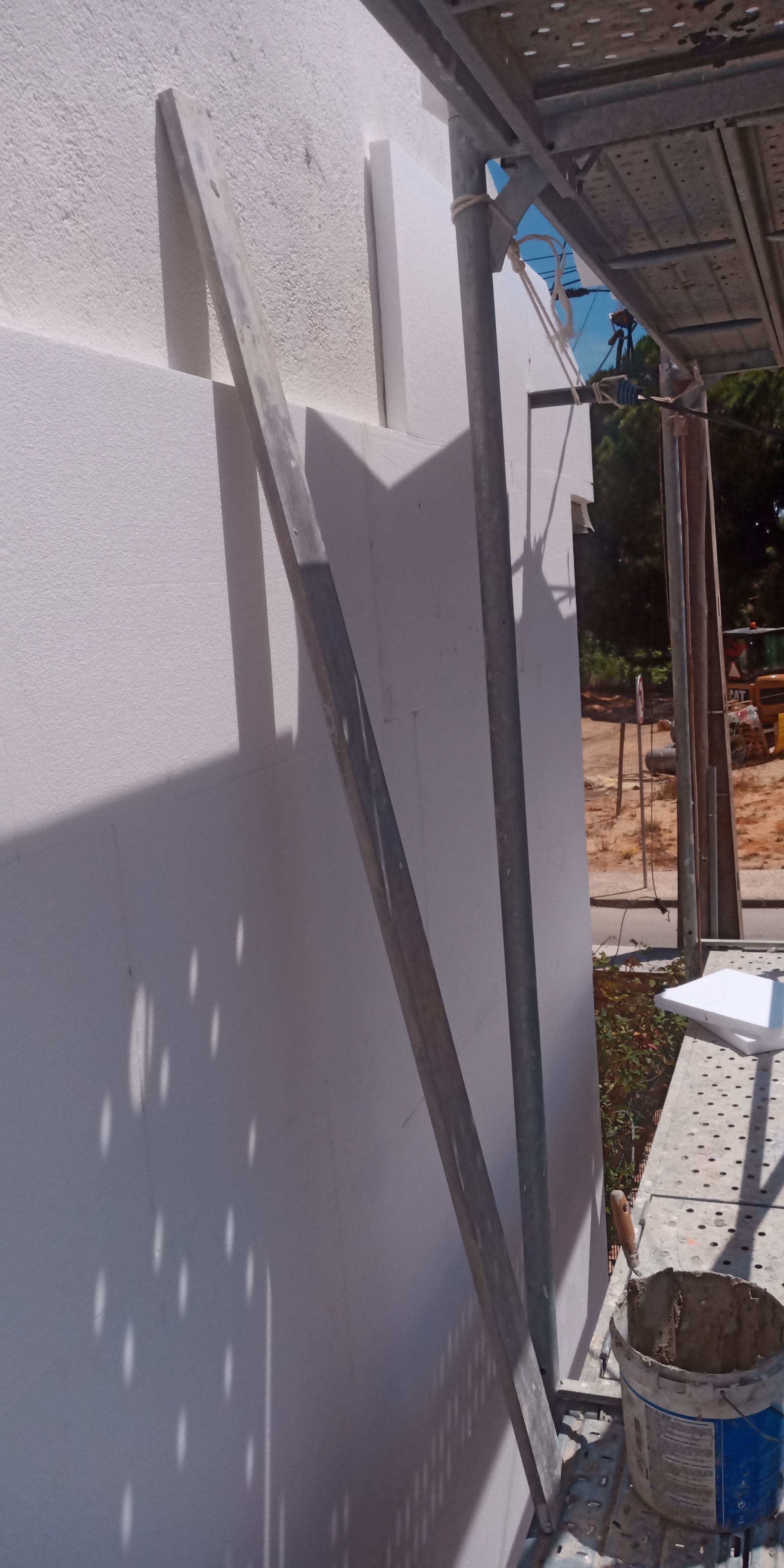 Parede externa rebocada de branco com régua metálica; canteiro de obras.