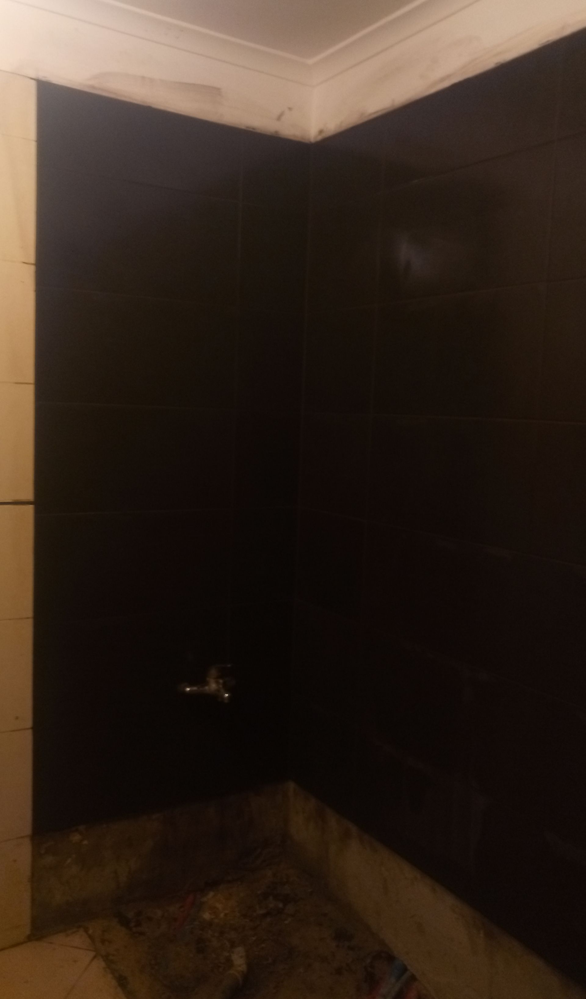 Canto de um banheiro com paredes revestidas de azulejos pretos, torneira prateada e piso inacabado.