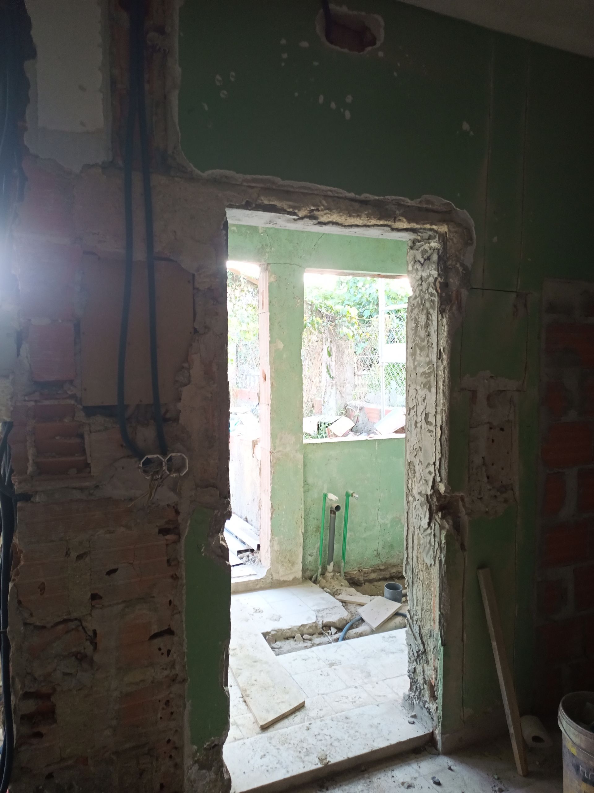 Canteiro de obras, porta em reforma. Paredes verdes, tijolos aparentes, fiação elétrica. A luz do sol entra pela porta do cômodo ao lado.