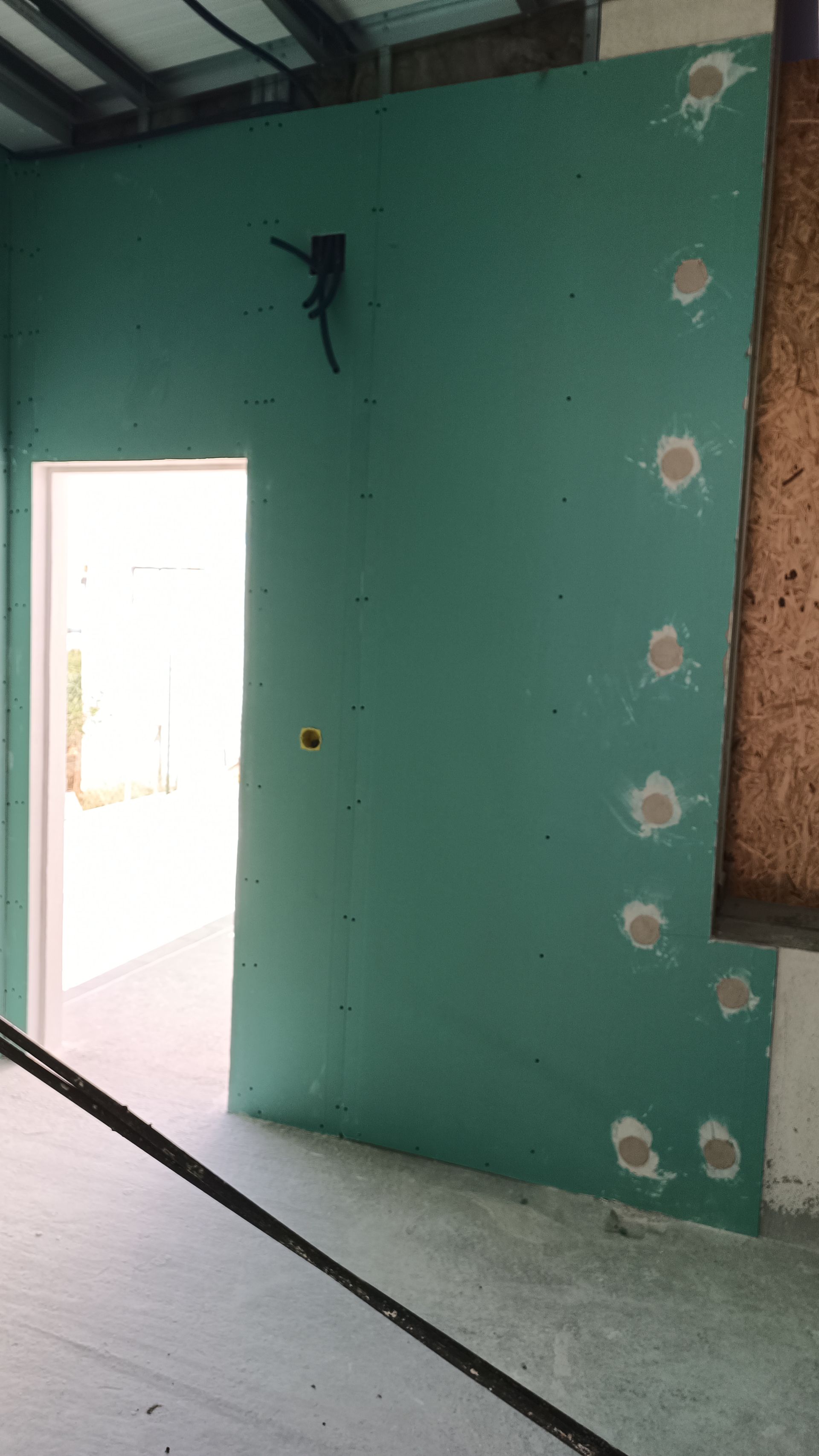 Parede de gesso cartonado verde com batente de porta, buracos preenchidos e fios elétricos.