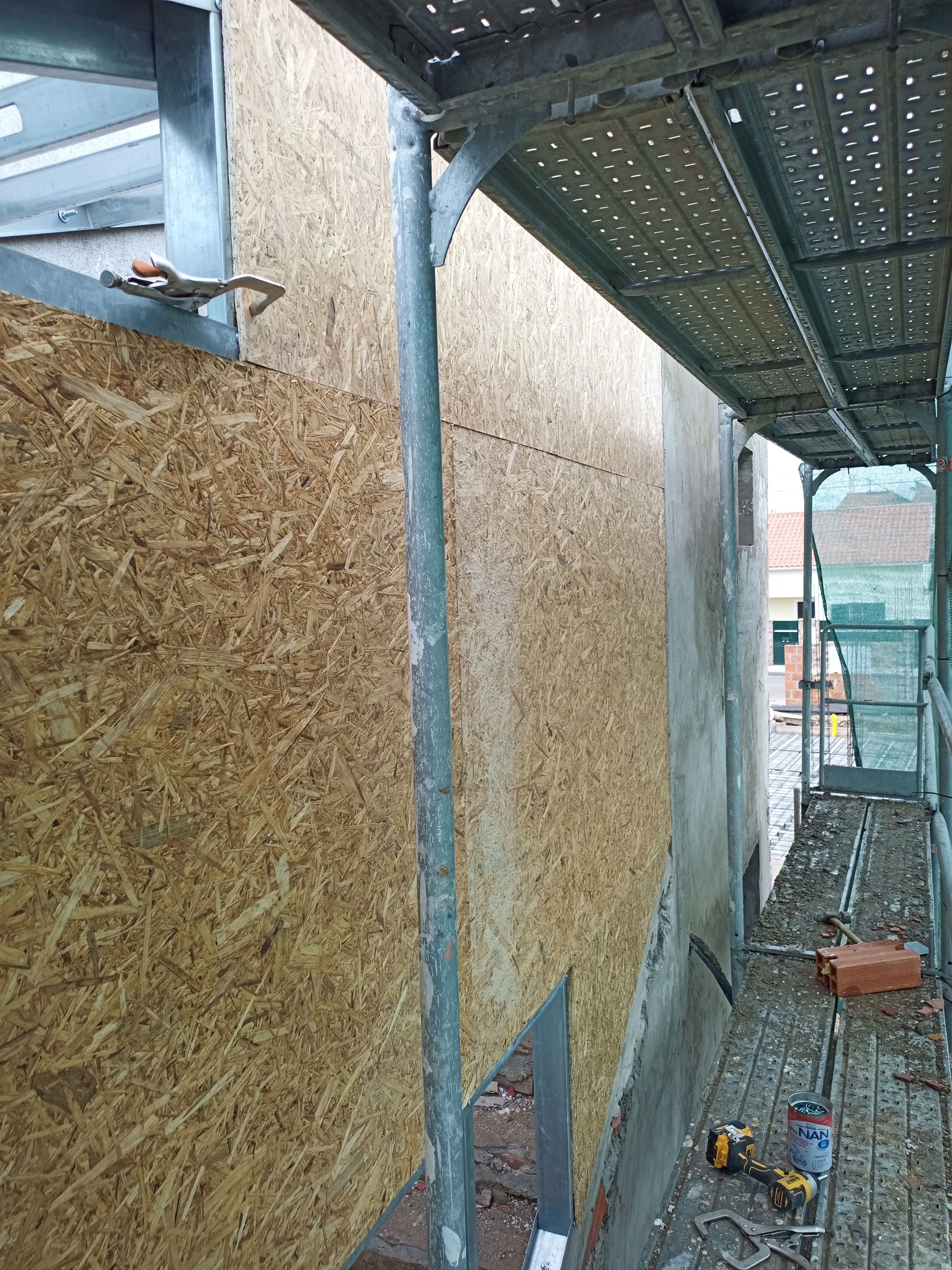 Painéis de OSB revestem a fachada de um edifício, ao lado de um andaime. Uma janela parcialmente emoldurada é visível.