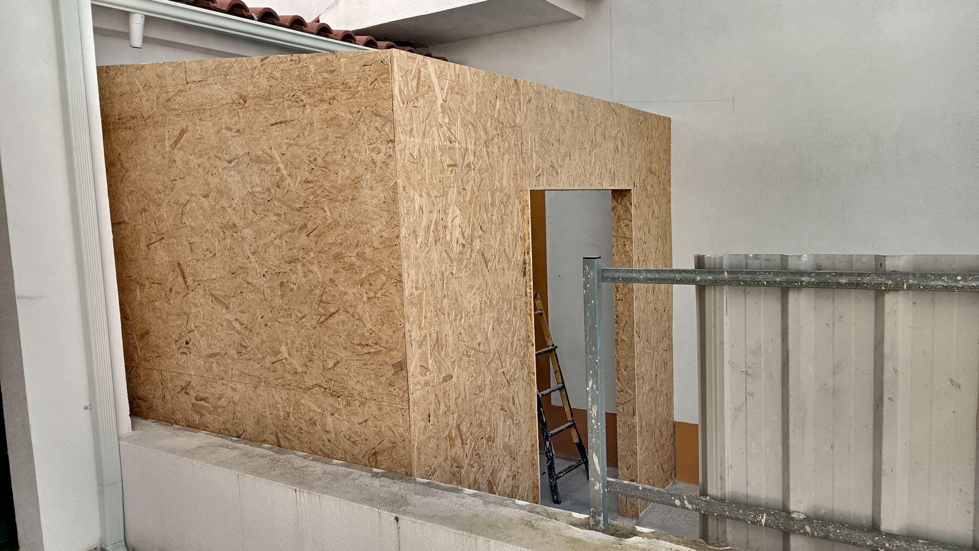 Estrutura de madeira em forma de caixa com porta, construída contra uma parede branca e uma cerca de metal.