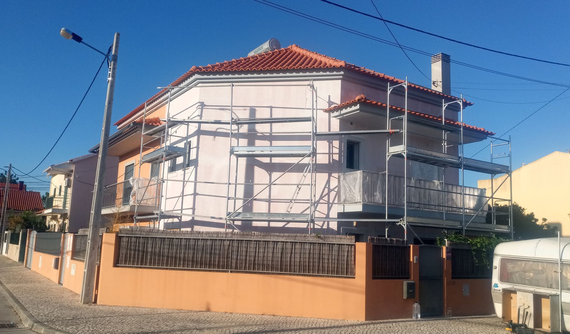 Casa com andaimes durante reforma externa; fachada laranja e branca, telhado de telha vermelha, céu limpo.