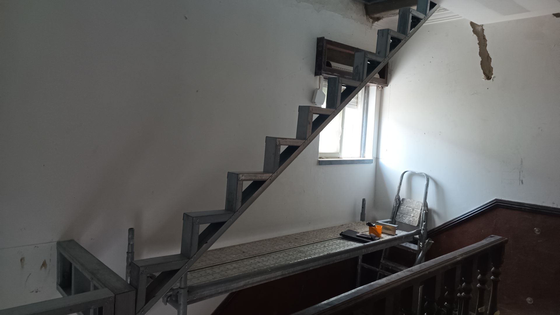 Escada metálica subindo uma parede branca, perto de uma janela, com corrimão visível no canto inferior direito.