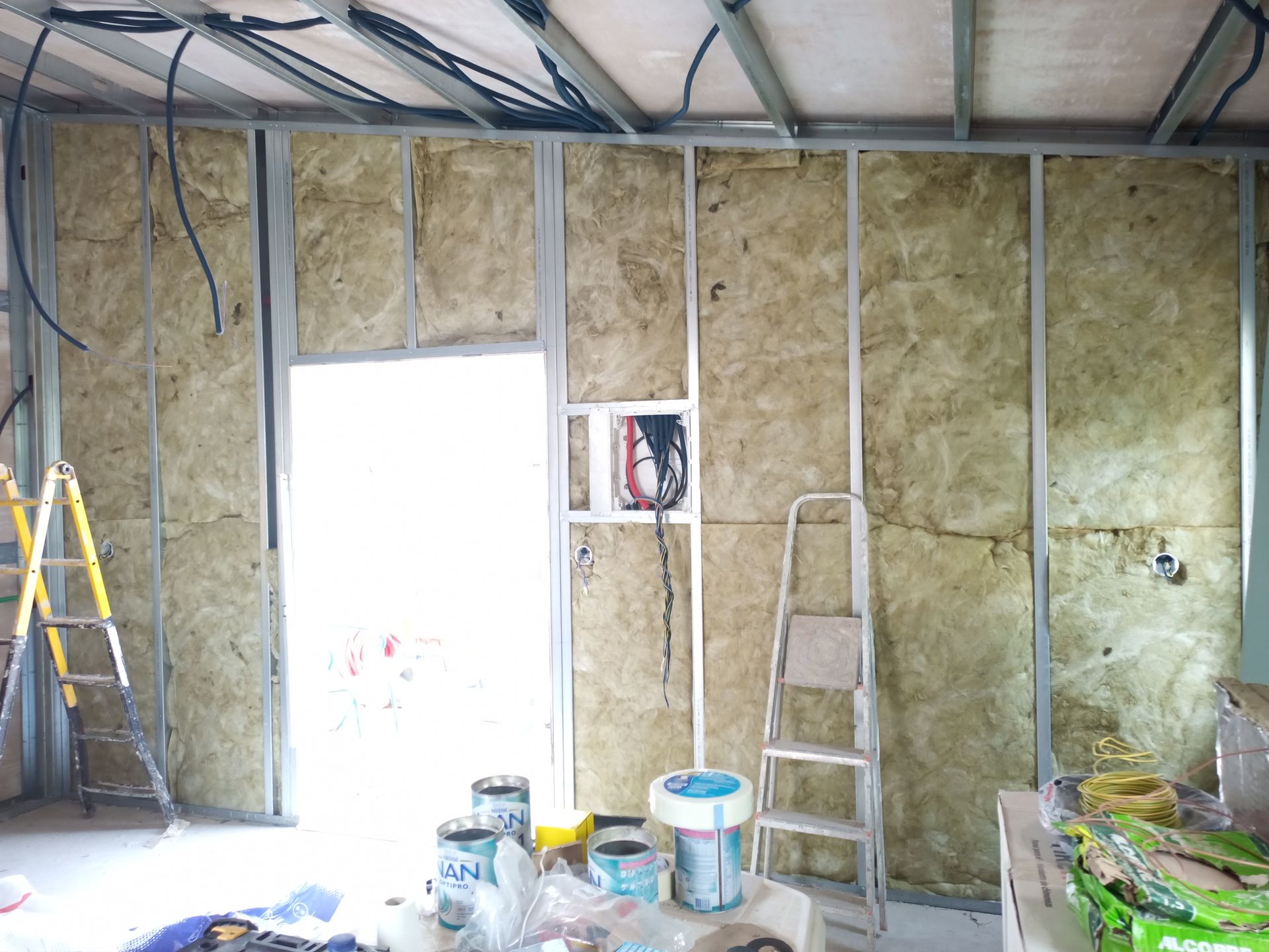 Parede interna em construção, estrutura metálica, isolamento instalado, fiação visível, escadas, latas de tinta no chão.