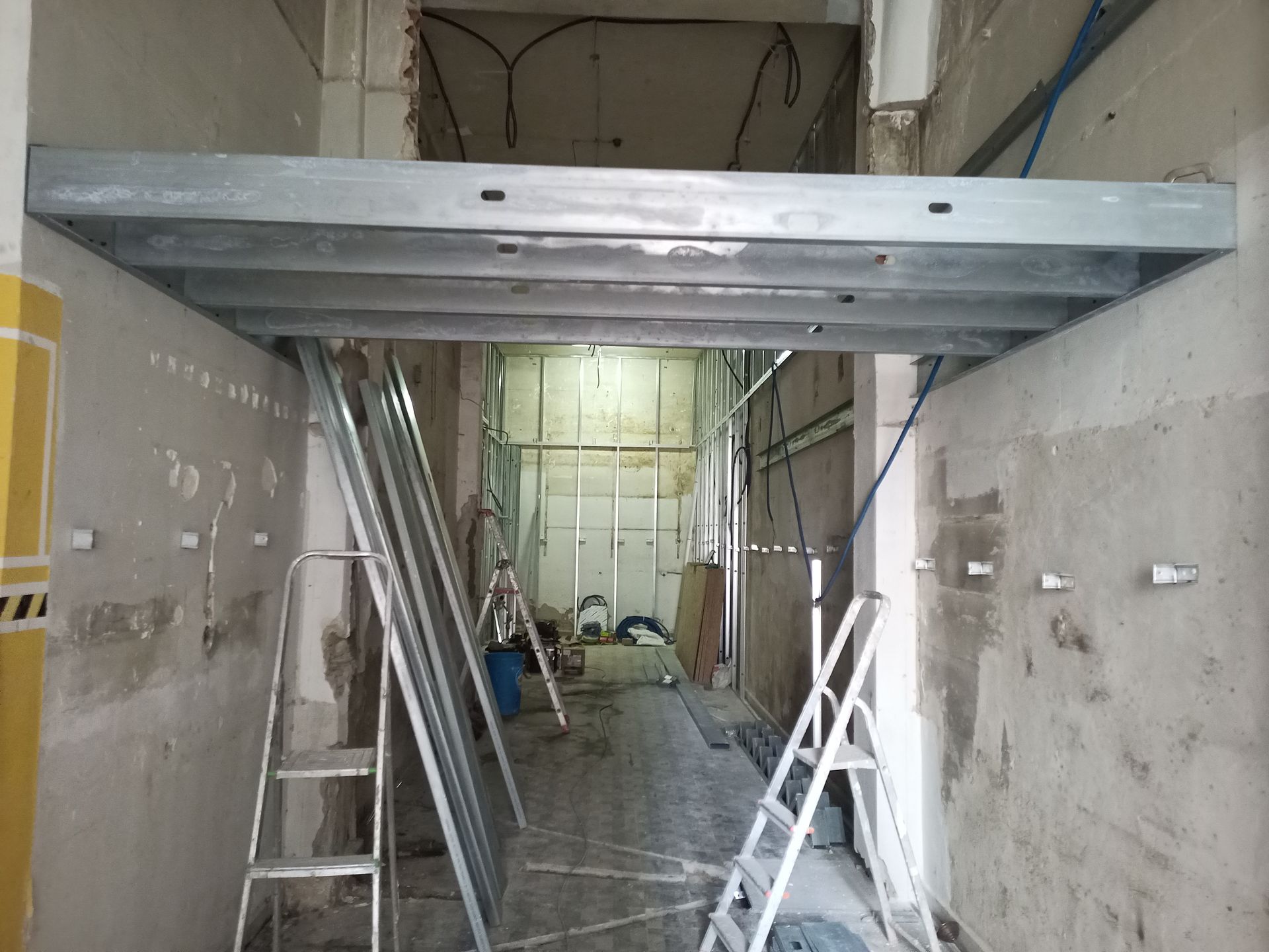 Canteiro de obras: Forro com estrutura metálica instalado em um corredor estreito com paredes expostas, escadas e materiais de construção.