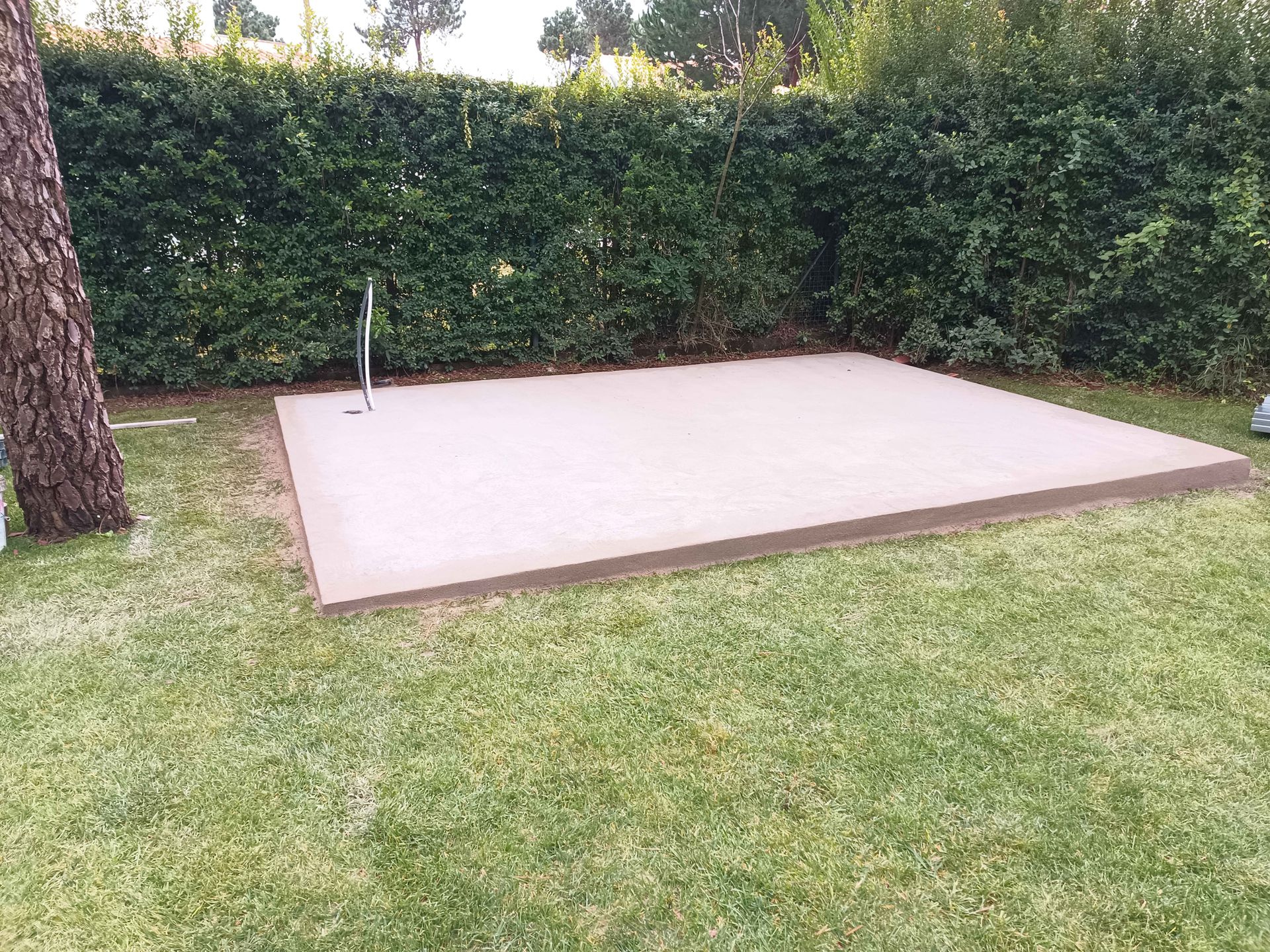 Uma plataforma retangular de concreto em um gramado, com uma cerca viva verde ao fundo e um poste de metal.