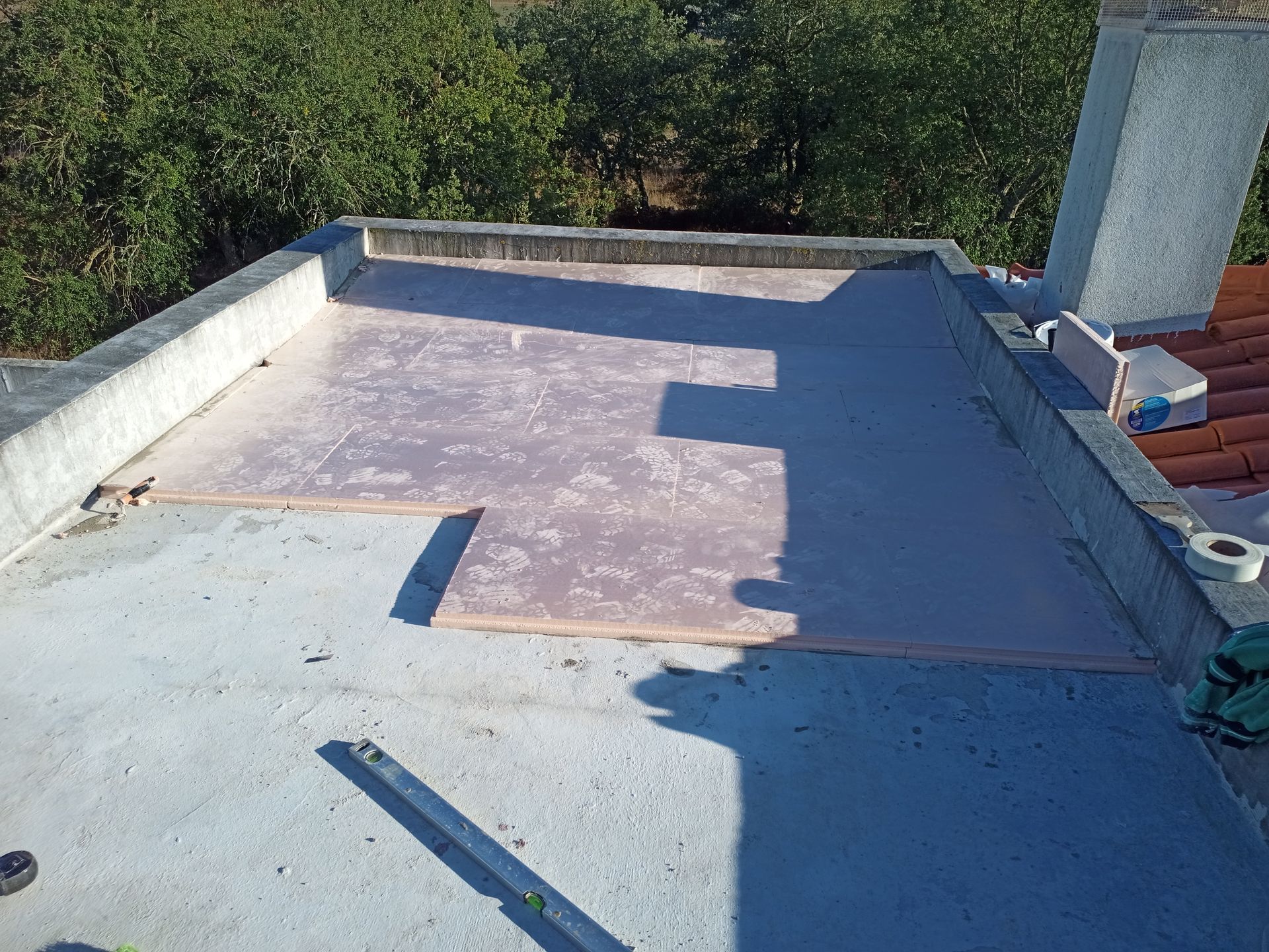 Telhado plano com painéis isolantes e borda de concreto ao redor, árvores ao fundo.