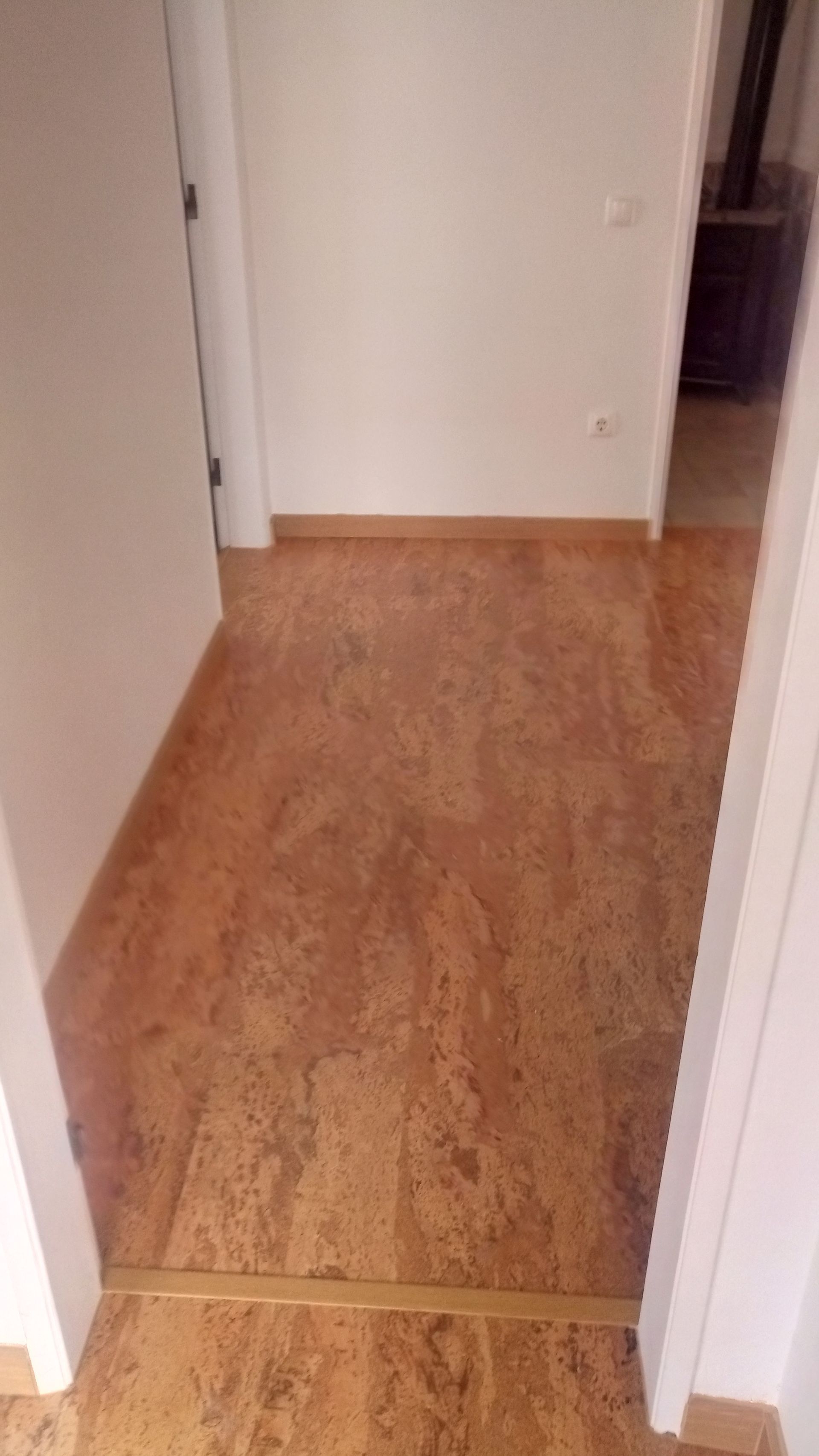 Piso de cortiça em um corredor que leva a outro cômodo. Portas e paredes brancas são visíveis.