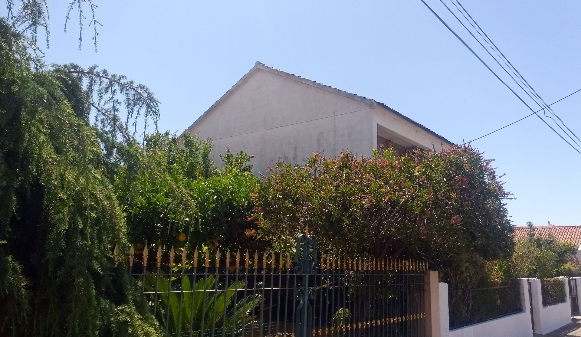 Casa branca com telhado de duas águas, parcialmente encoberta por arbustos verdes e uma cerca preta sob um céu claro.