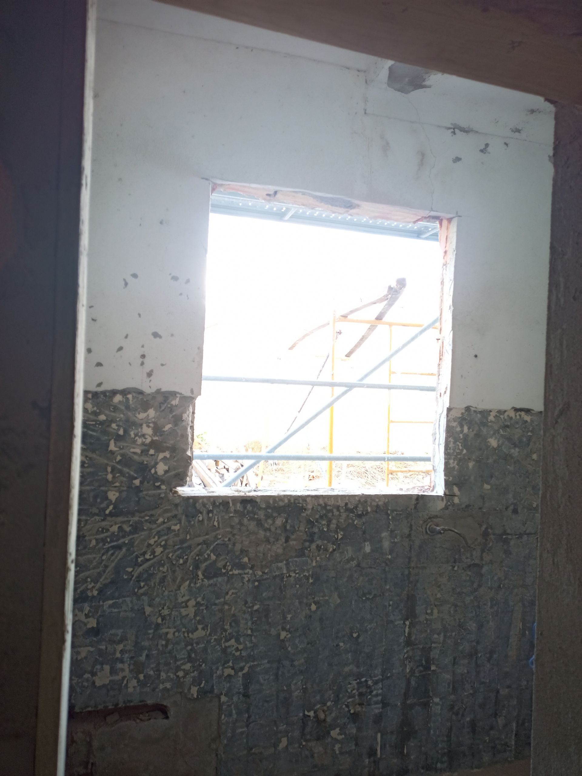 Abertura de janela em uma parede, coberta com tinta texturizada, com luz solar intensa e andaimes visíveis através da janela.