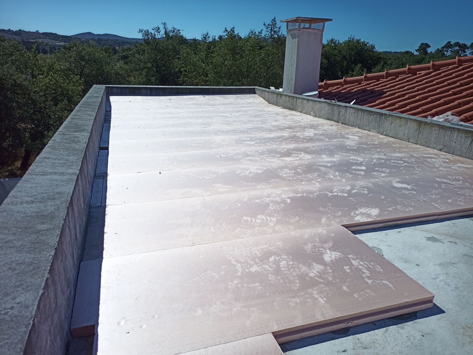 Telhado plano com painéis de isolamento rosa parcialmente instalados, chaminé e árvores ao fundo.