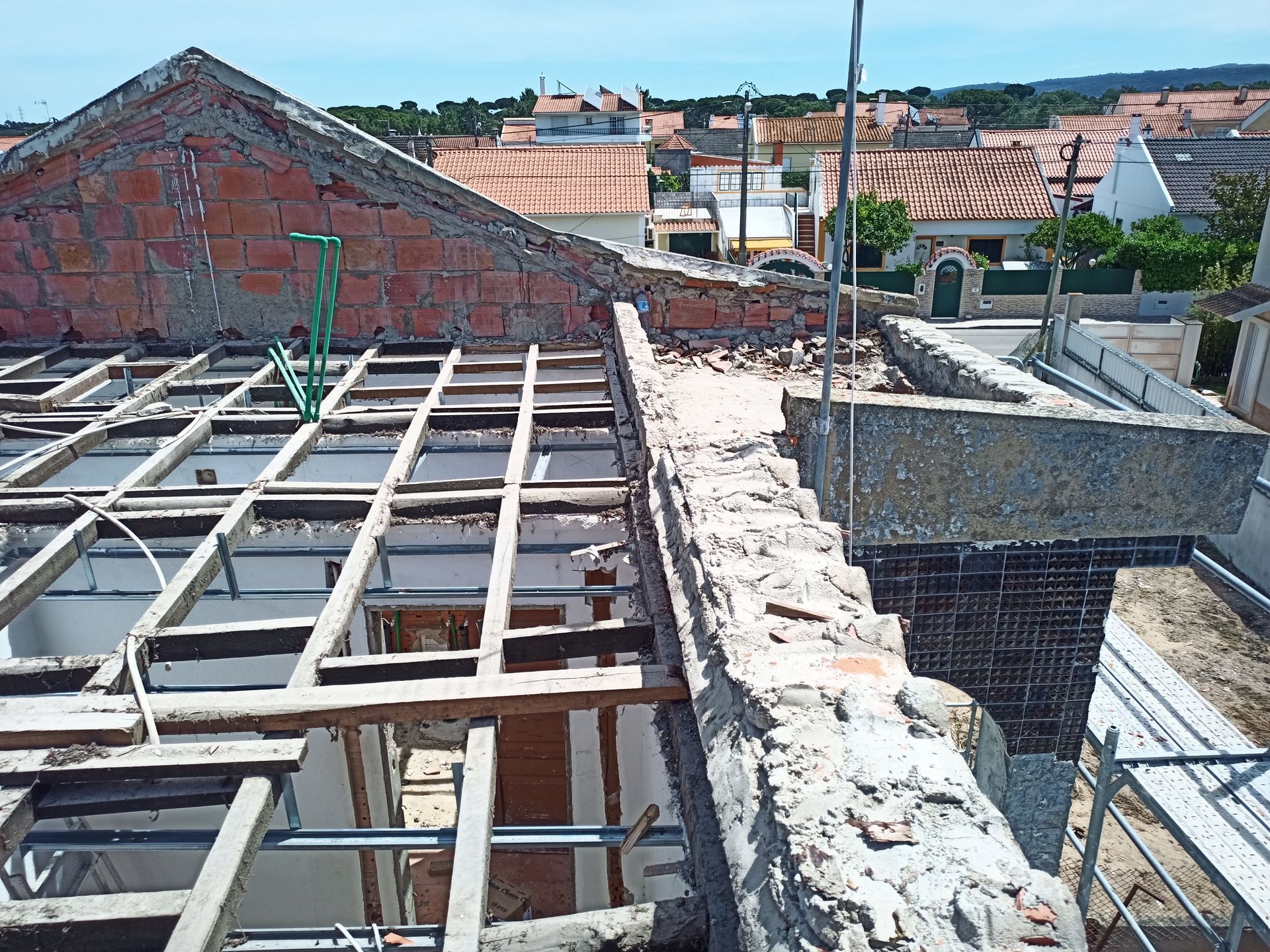 Canteiro de obras com telhado aparente, vigas e alvenaria expostas, e vista para a cidade.