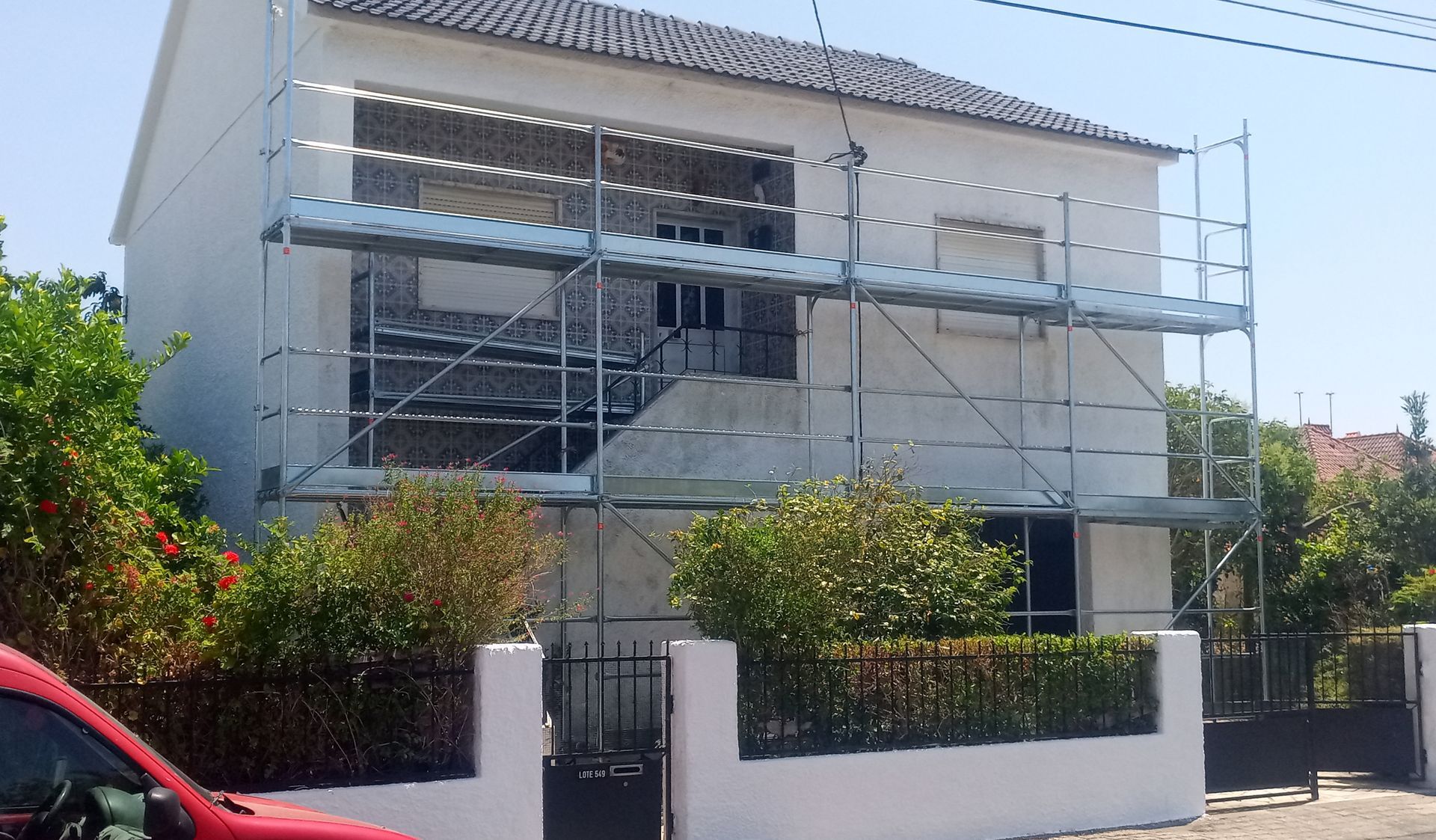Andaimes em uma casa branca de dois andares, em reforma. Arbustos verdes e um carro vermelho estão em primeiro plano.