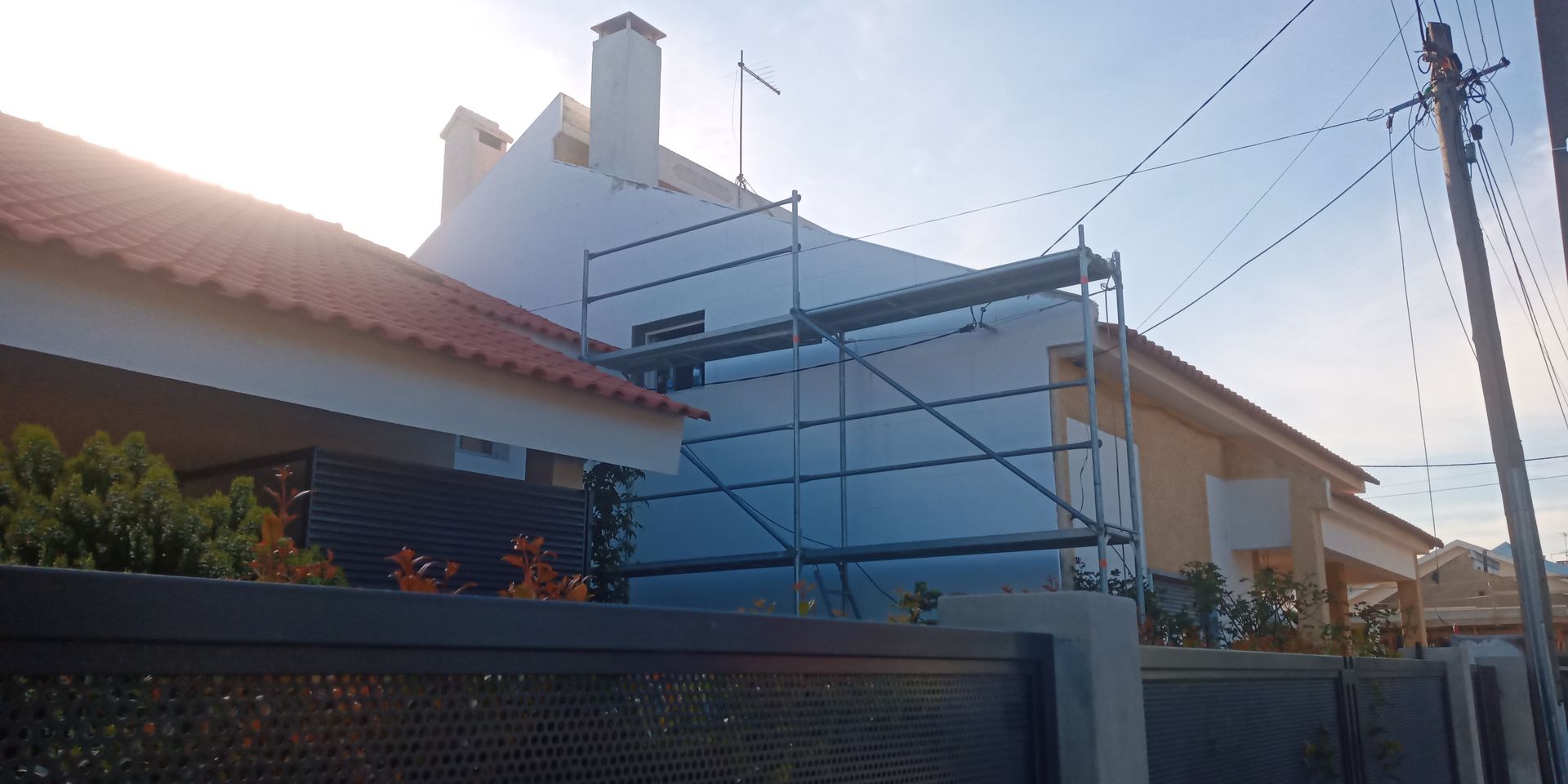 Andaimes encostados em uma casa branca; uma cerca e um poste de luz estão em primeiro plano.