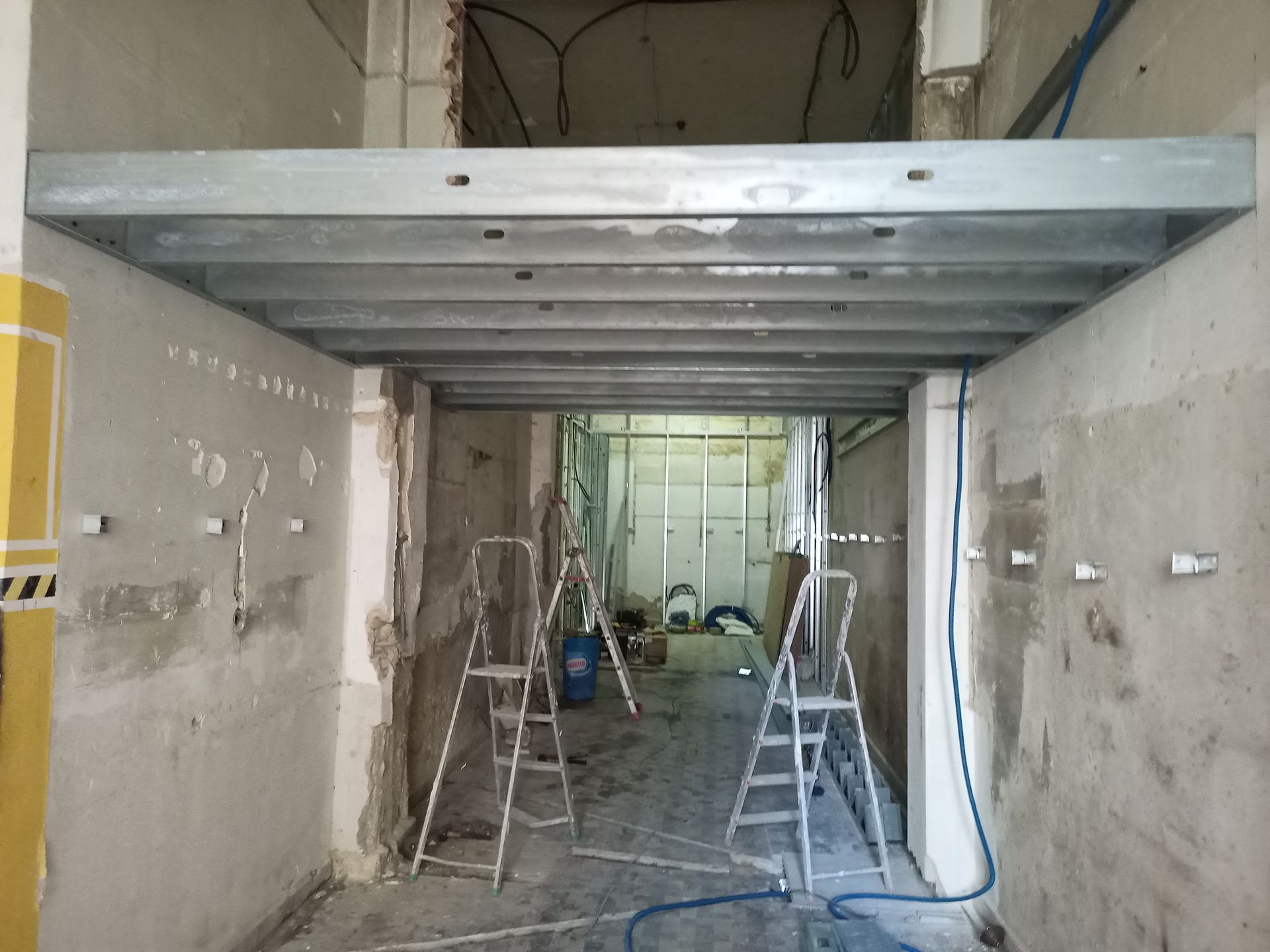 Construção de teto com estrutura metálica em um corredor estreito durante uma reforma, com escadas e ferramentas visíveis.