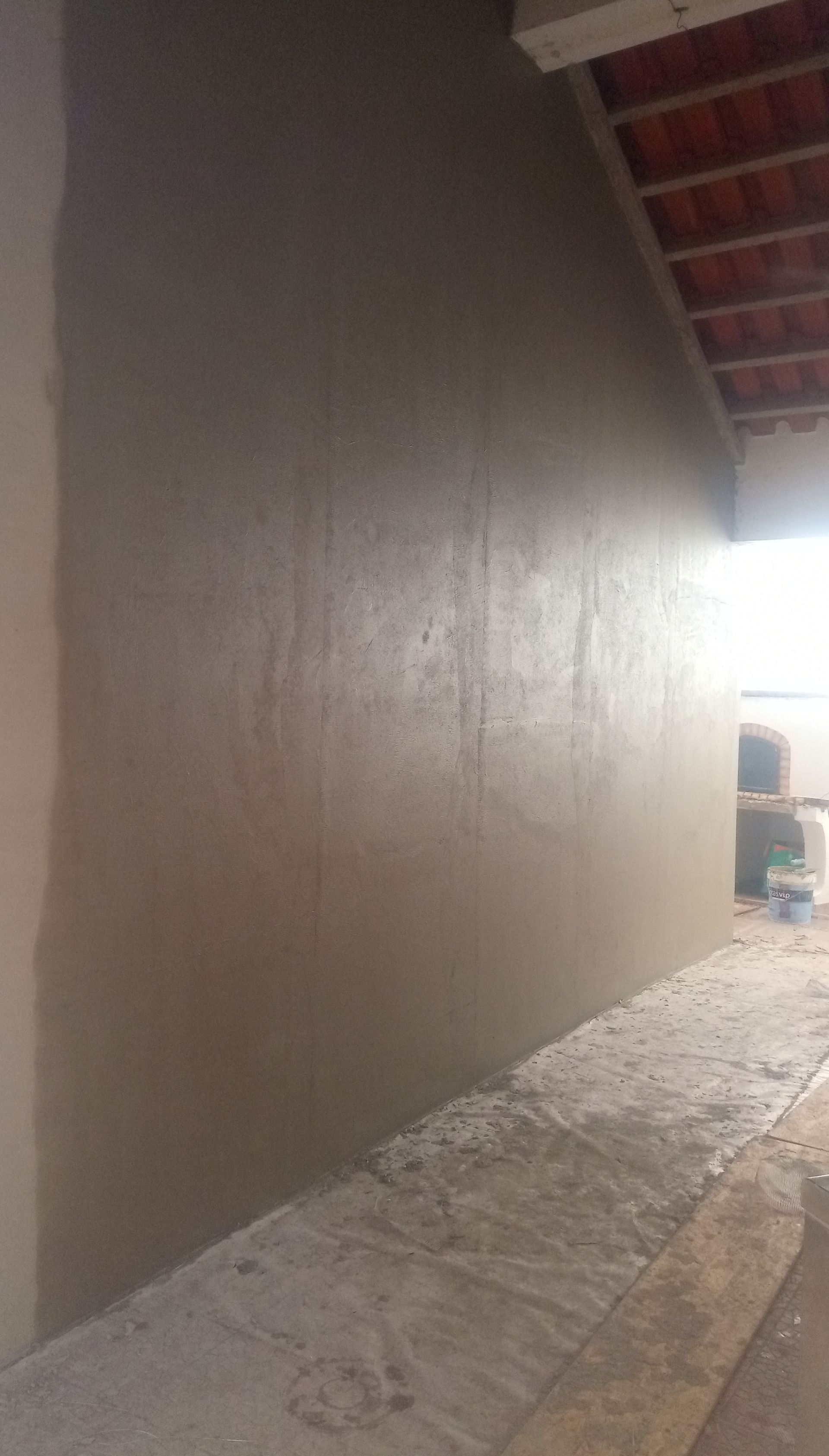 Parede com reboco cinza em processo de aplicação, com calçada de concreto inacabada e telhado de telhas vermelhas visíveis.