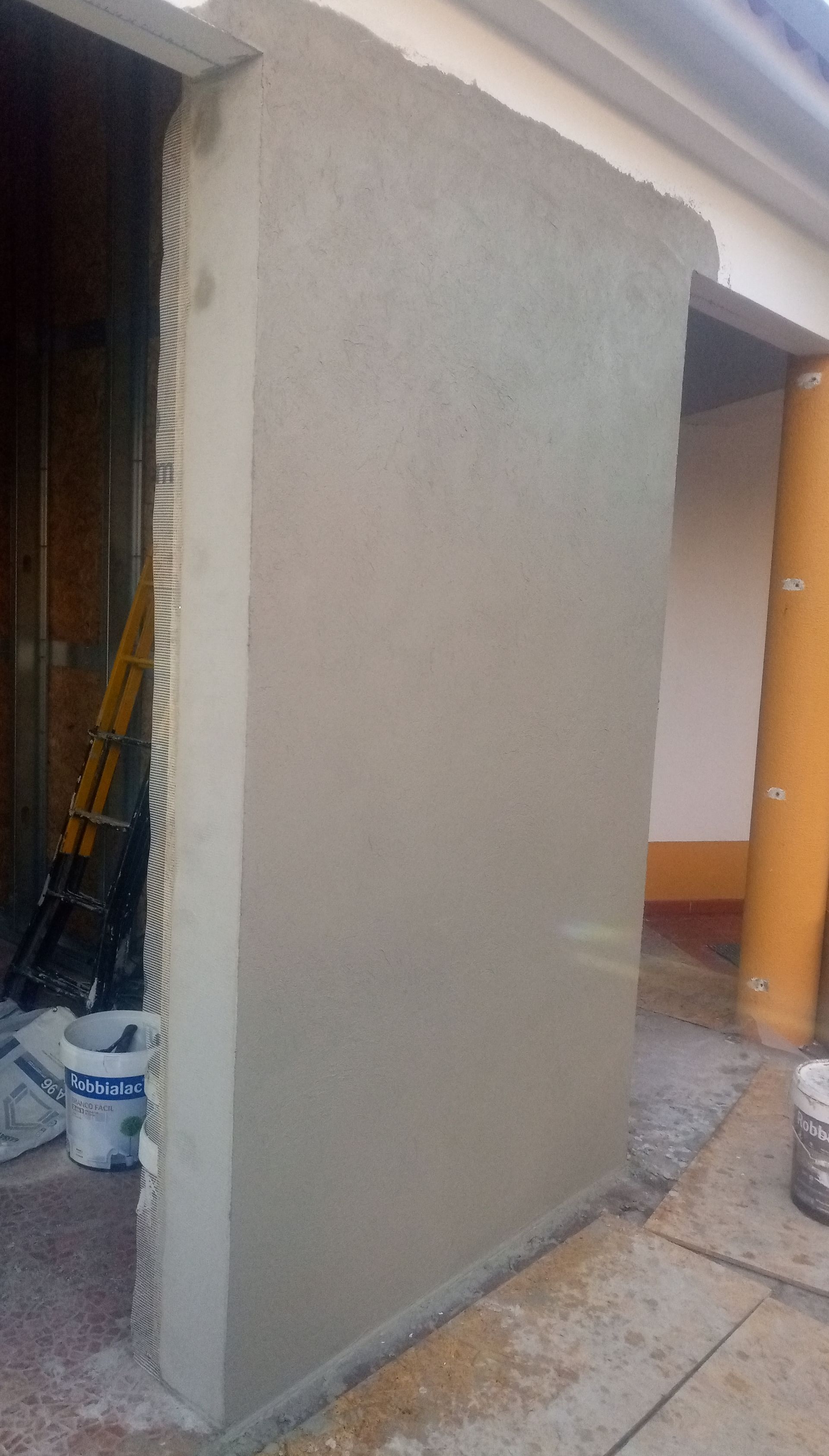 Parede parcialmente rebocada na parte externa de um edifício. A parede é cinza-clara com superfície texturizada e é possível ver o batente de uma porta.