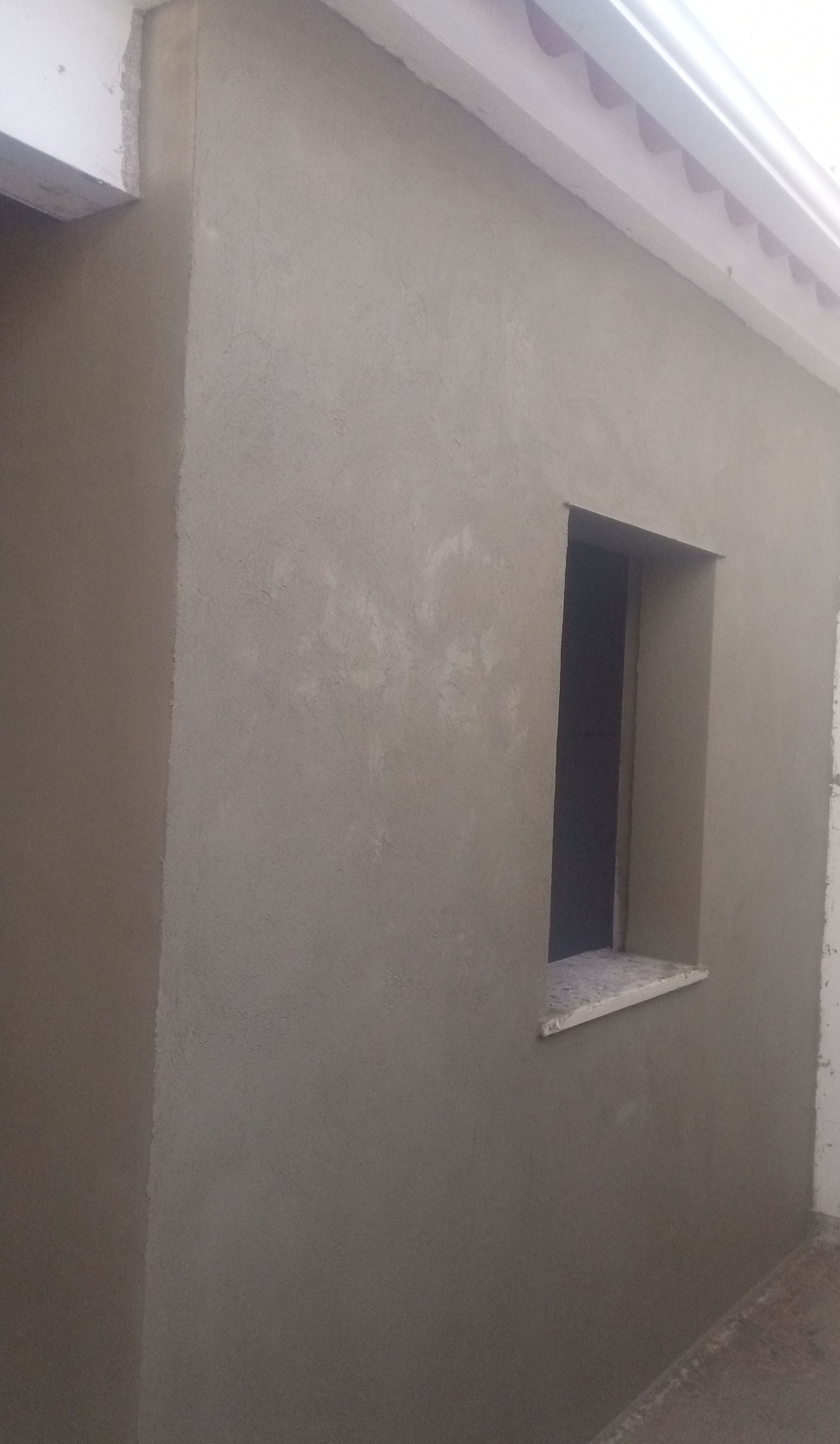 Parede externa com uma pequena janela, revestida de estuque cinza. A parede está parcialmente na sombra.
