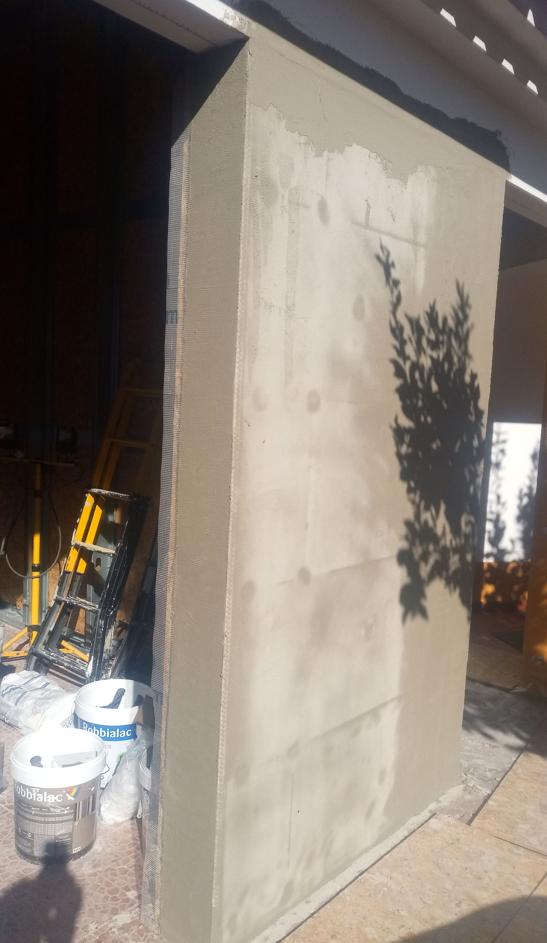 Um pilar retangular recém-rebocado do lado de fora. O pilar é cinza claro e está em sombra parcial. Escadas e baldes estão próximos.
