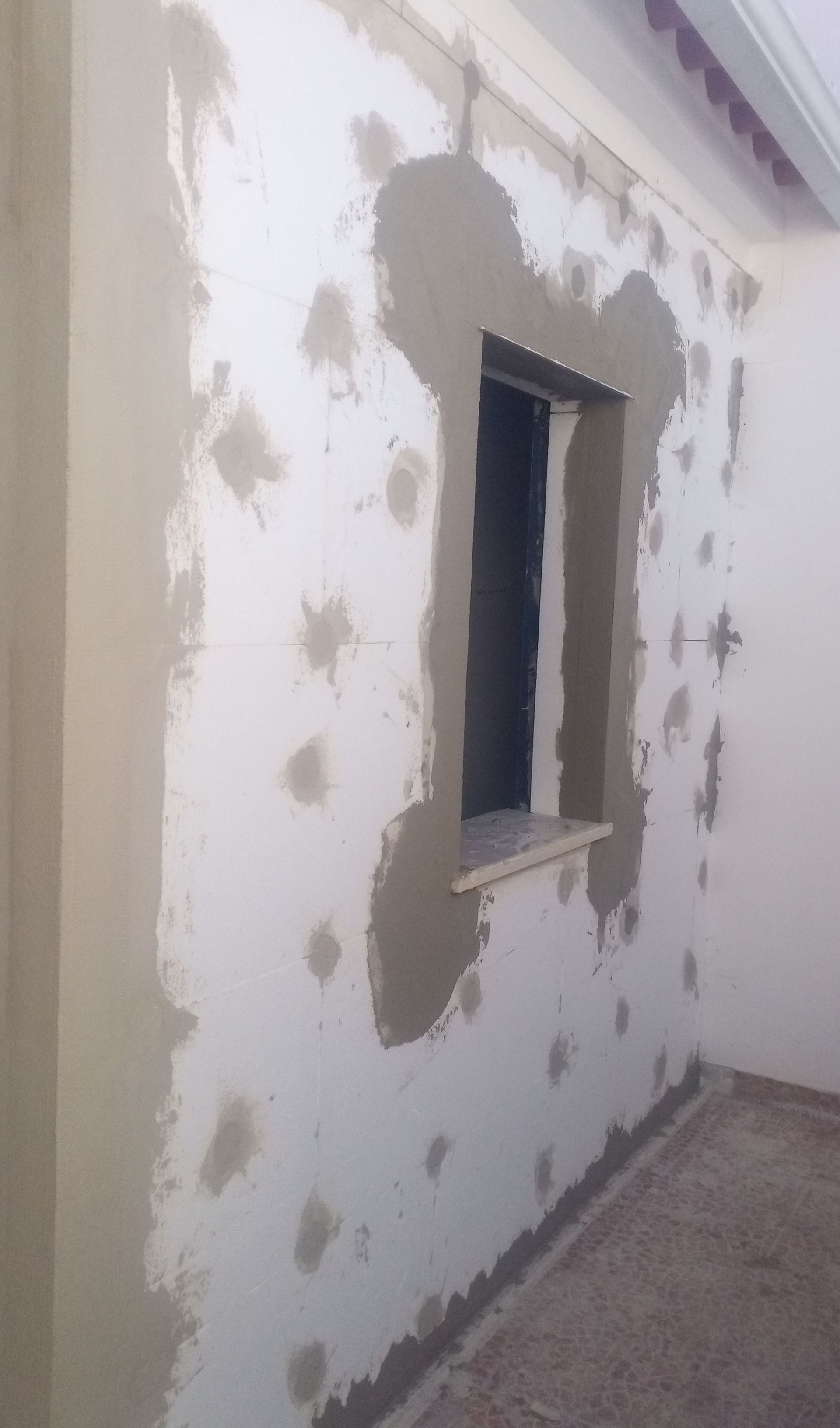 Parede exterior com janela, parcialmente rebocada com argamassa cinza clara, remendos e moldura.