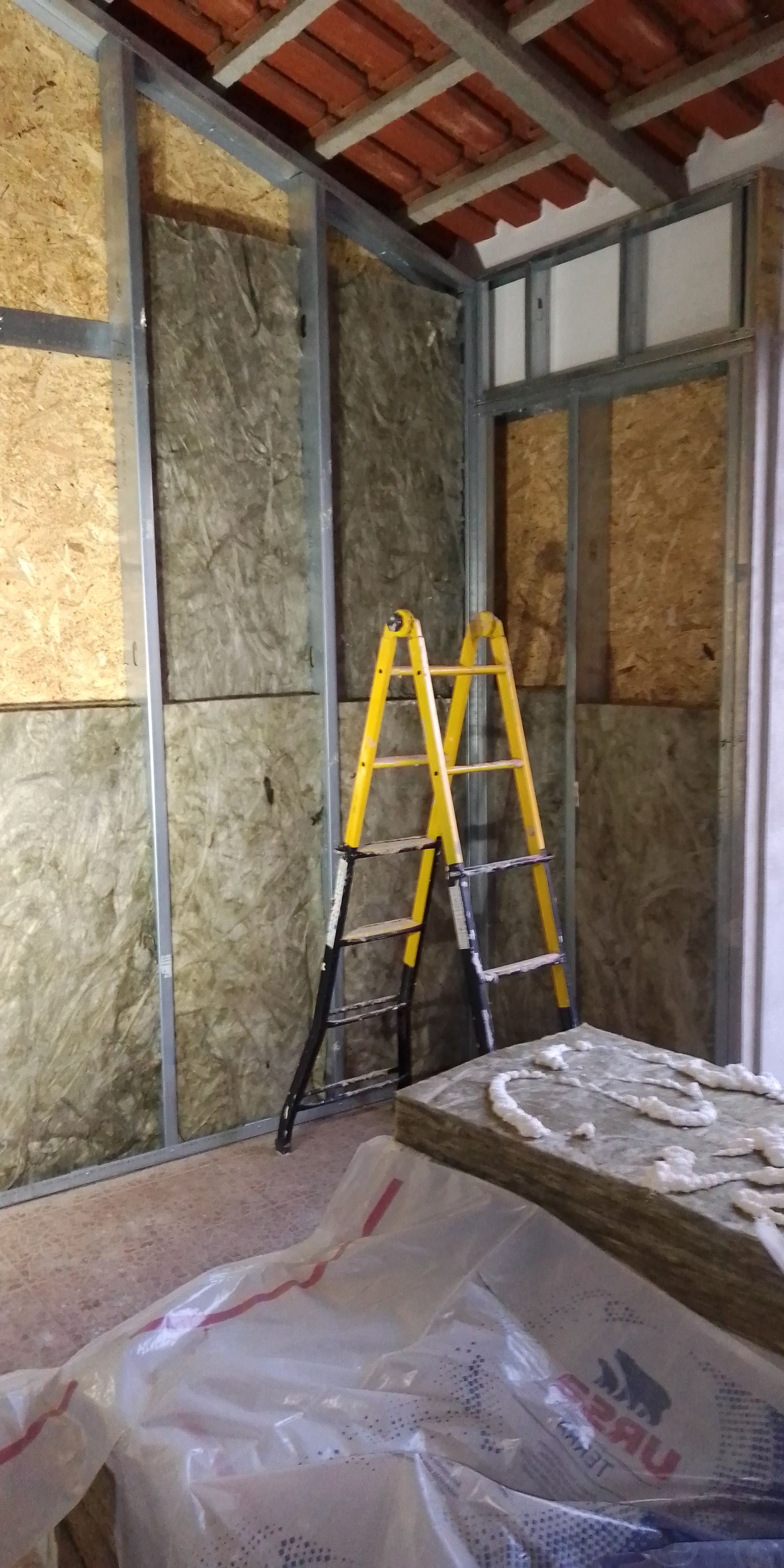 Construção interna em andamento, com estrutura de aço, isolamento e uma escada amarela.