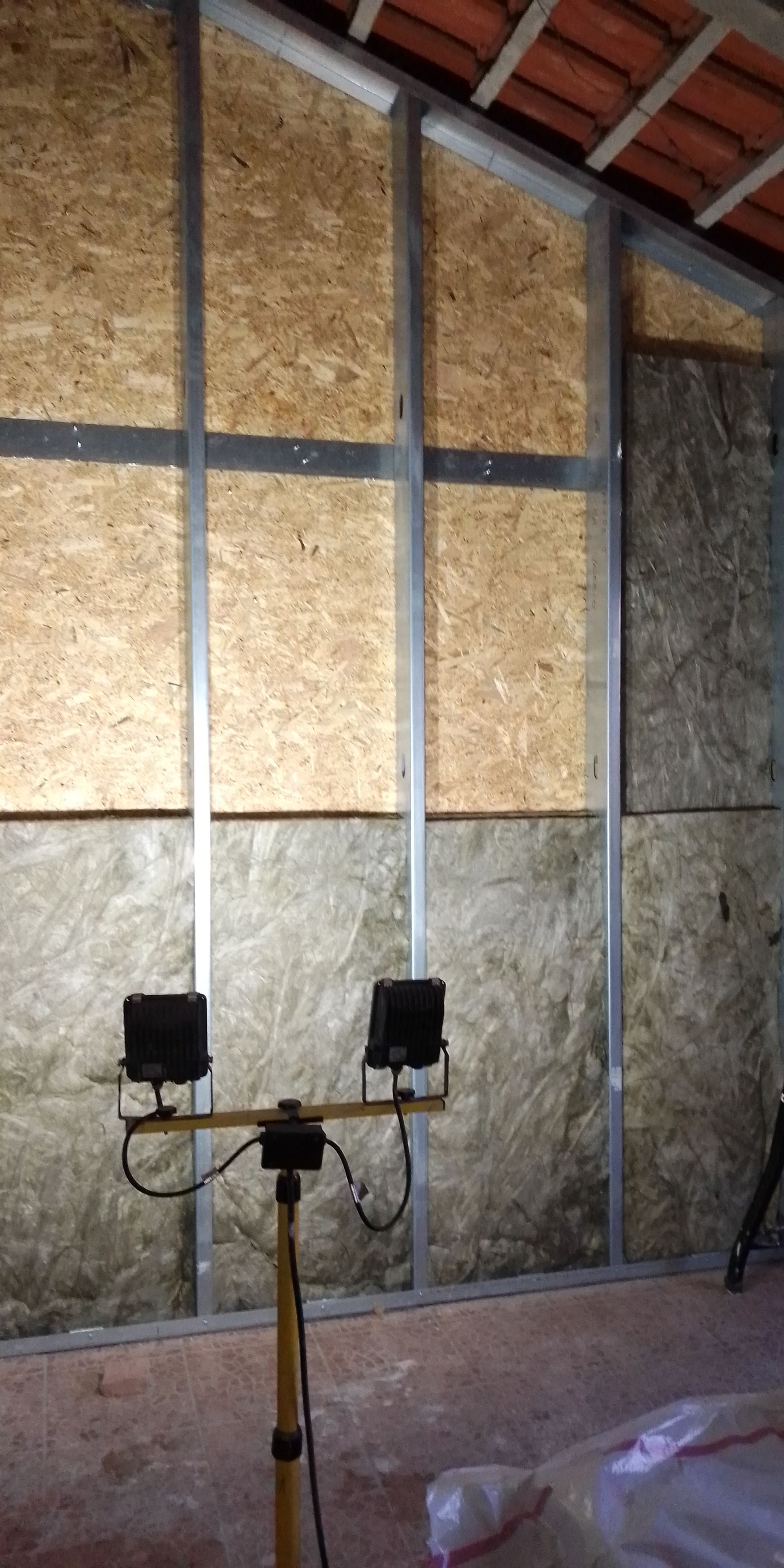 Parede parcialmente construída com isolamento, estrutura metálica e uma luminária de trabalho. Vigas de madeira visíveis.