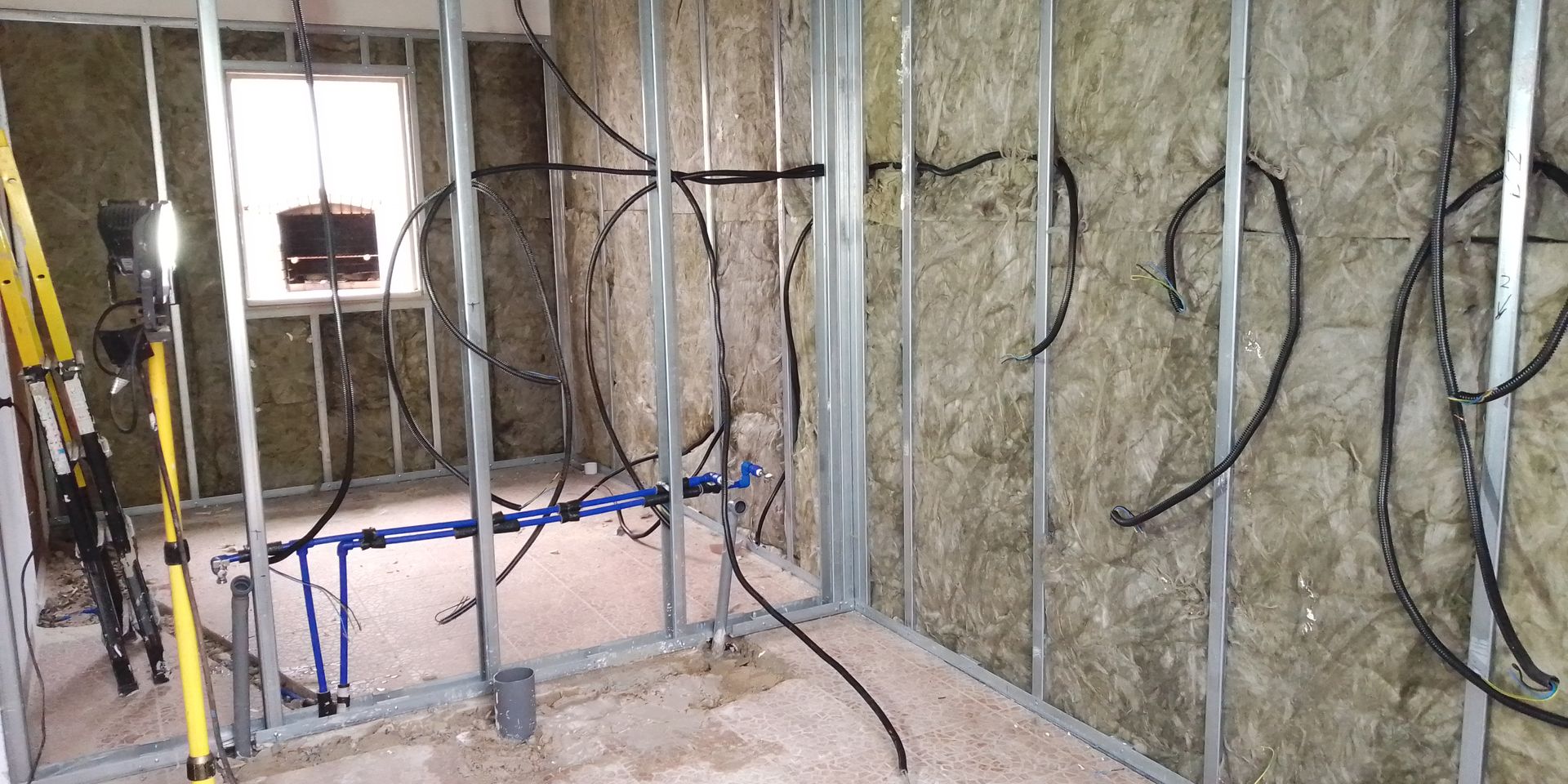 Vista interna de uma sala em construção com estrutura metálica, fiação e isolamento.