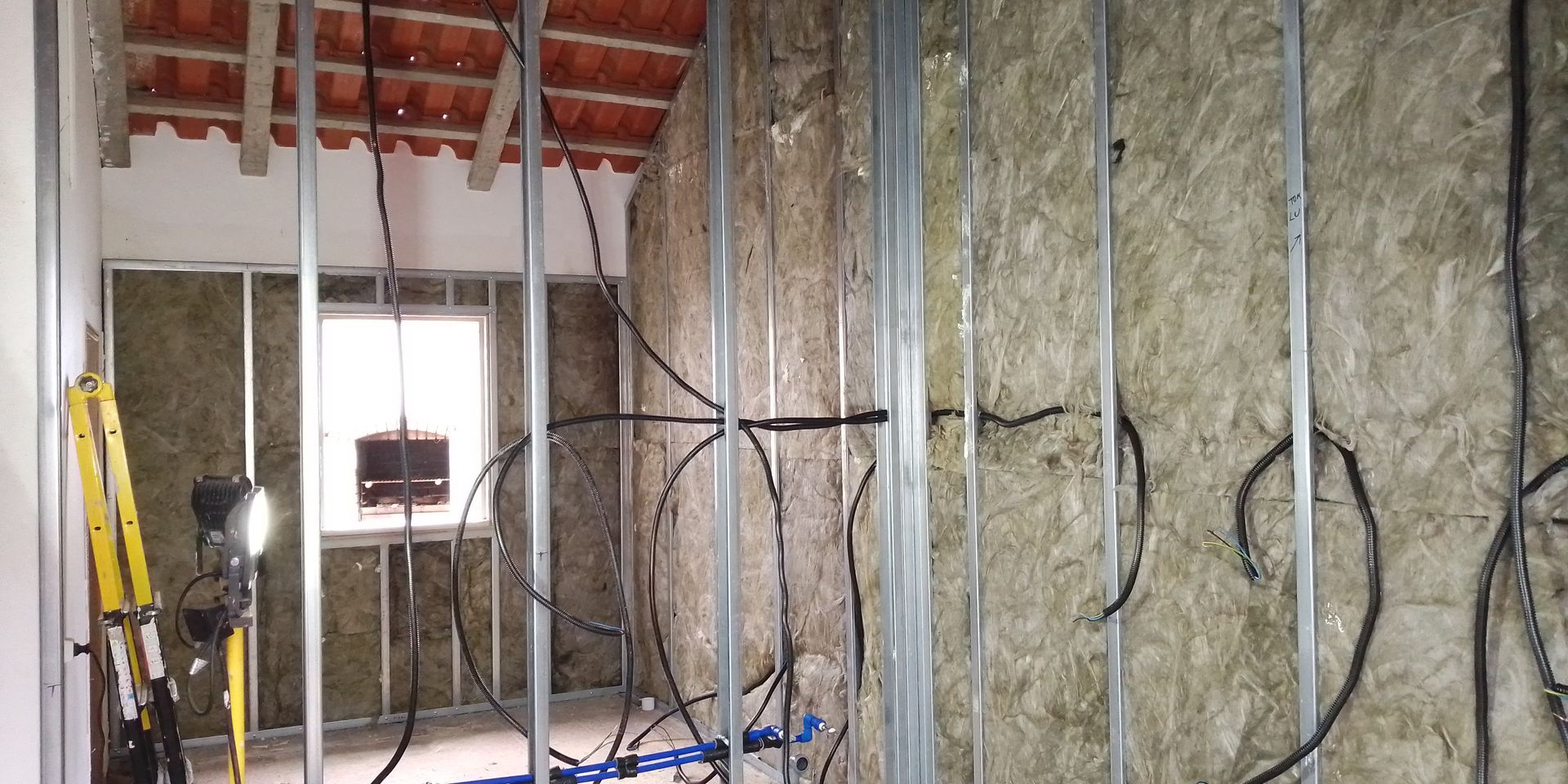 Construção interna em andamento, com estrutura metálica, isolamento, fiação elétrica e uma janela visíveis.