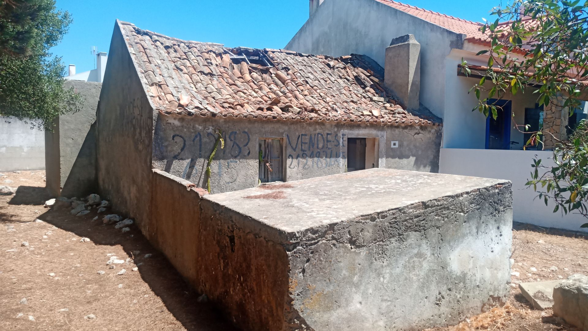 Prédio de concreto dilapidado com telhado de telhas, grafites e porta pequena. Ambiente externo ensolarado.