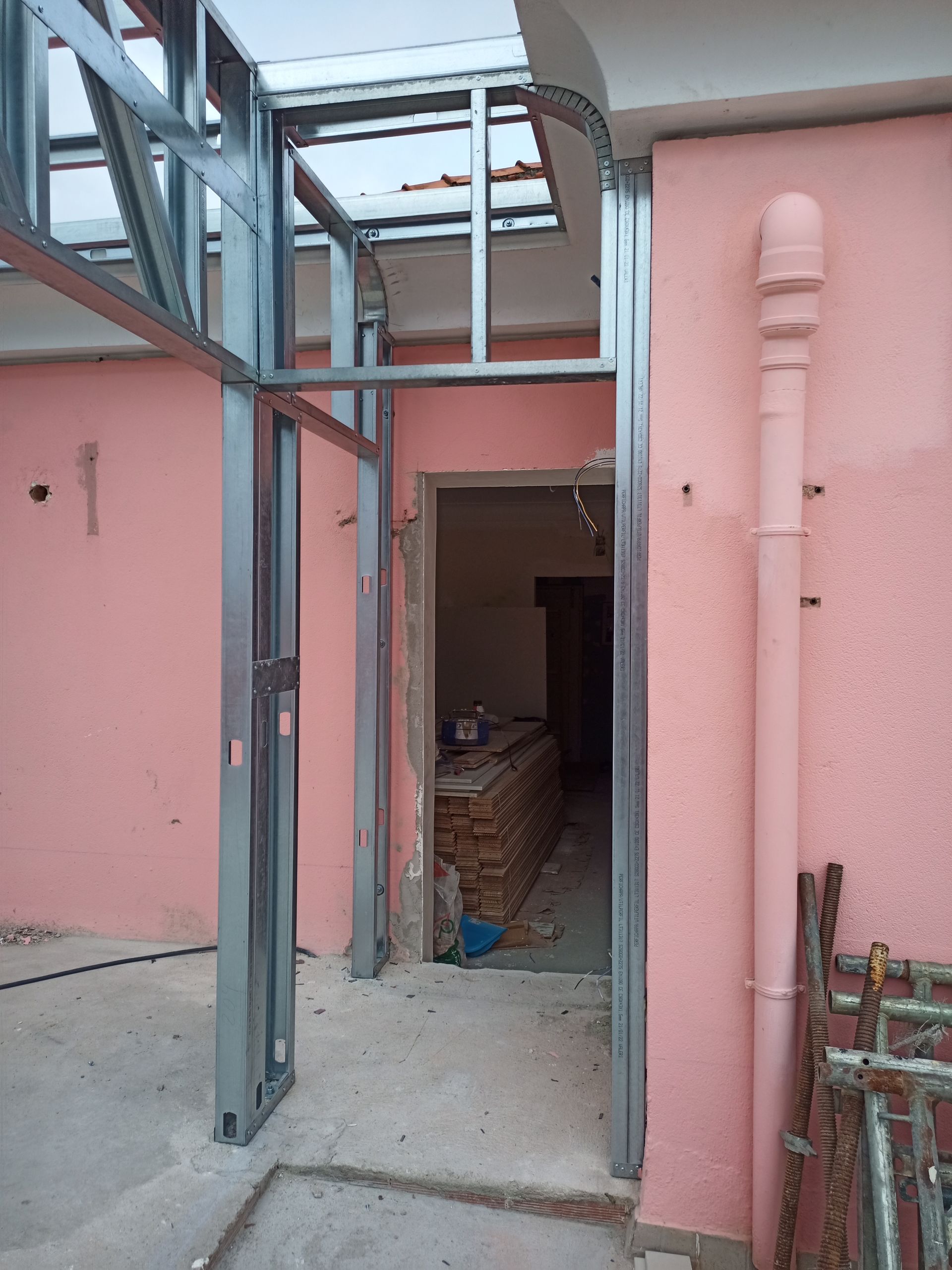 Estrutura metálica em torno de uma entrada, contra uma parede rosa. Um cano branco e alguns andaimes são visíveis.