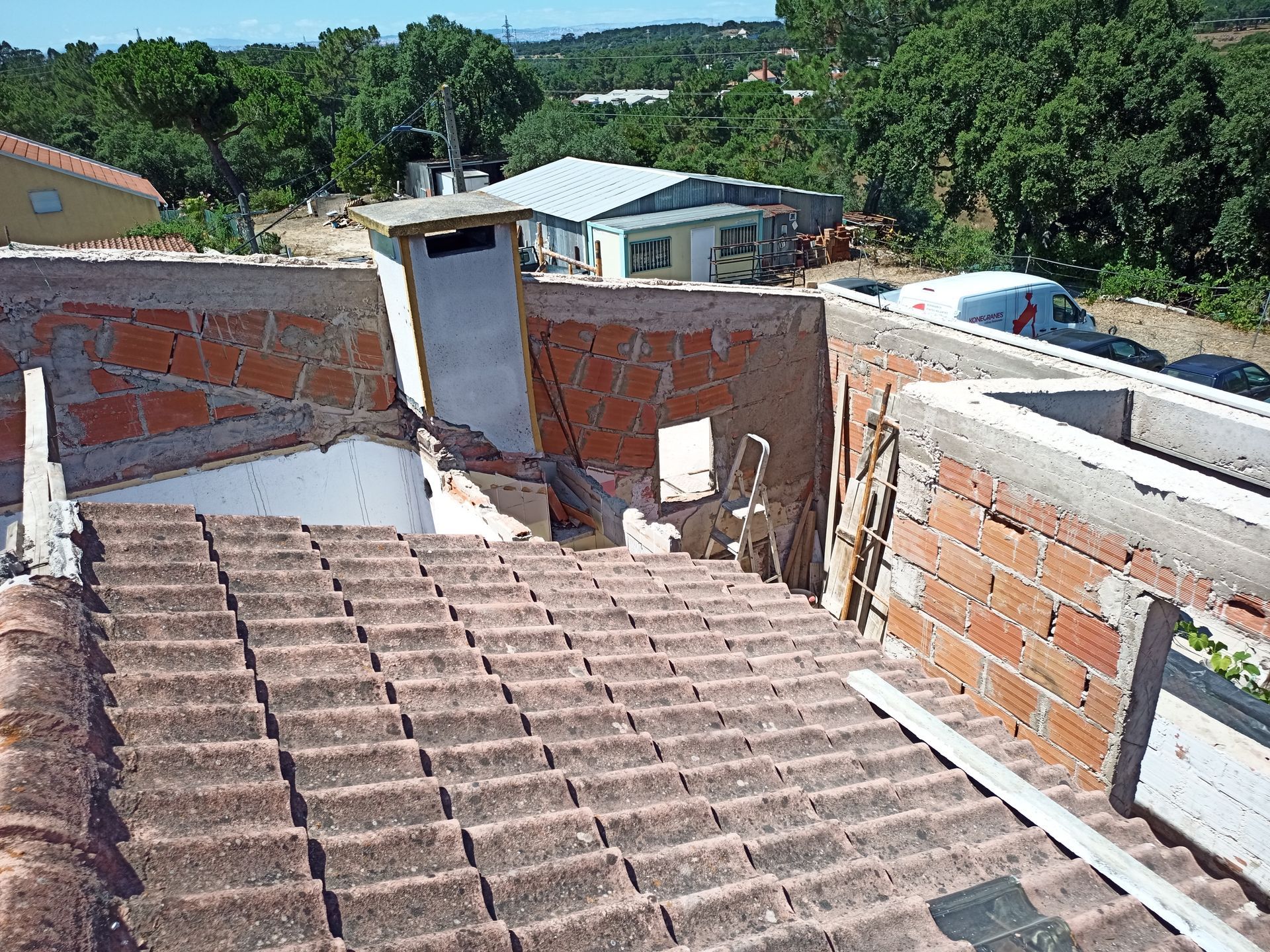 Telhado com estrutura de tijolos parcialmente desabada, tendo como pano de fundo árvores e edifícios em um dia ensolarado.