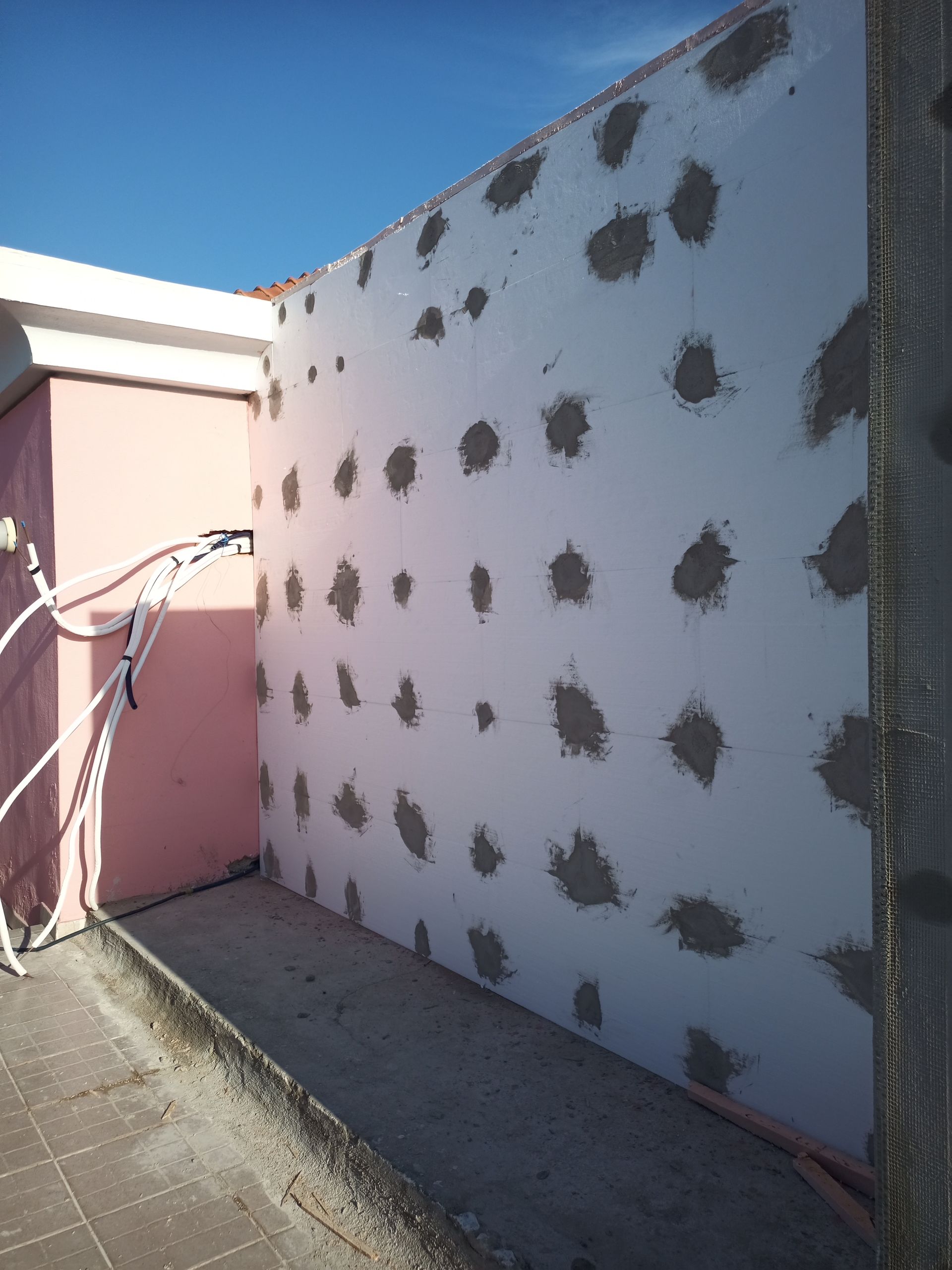 Parede branca com manchas cinzentas contra um céu azul, ao lado de uma parede rosa.