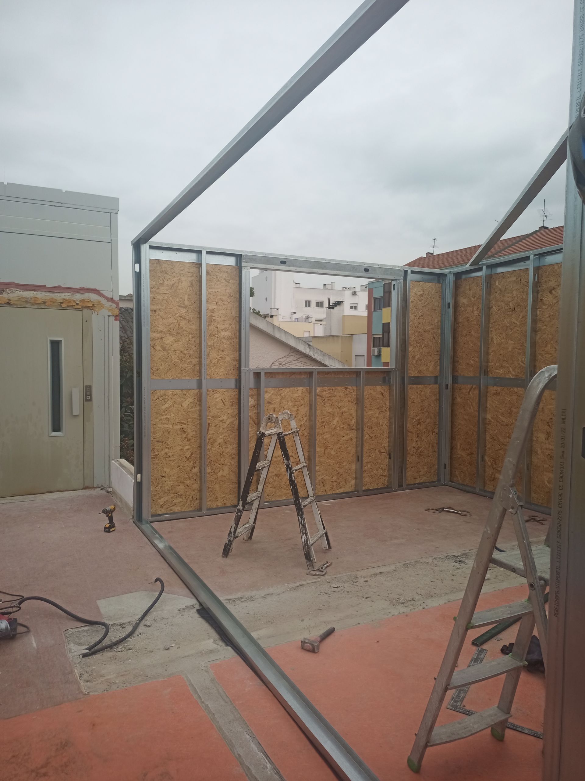 Construção de uma estrutura de cômodo com montantes metálicos e revestimento de OSB em área externa, com duas escadas presentes, céu nublado.