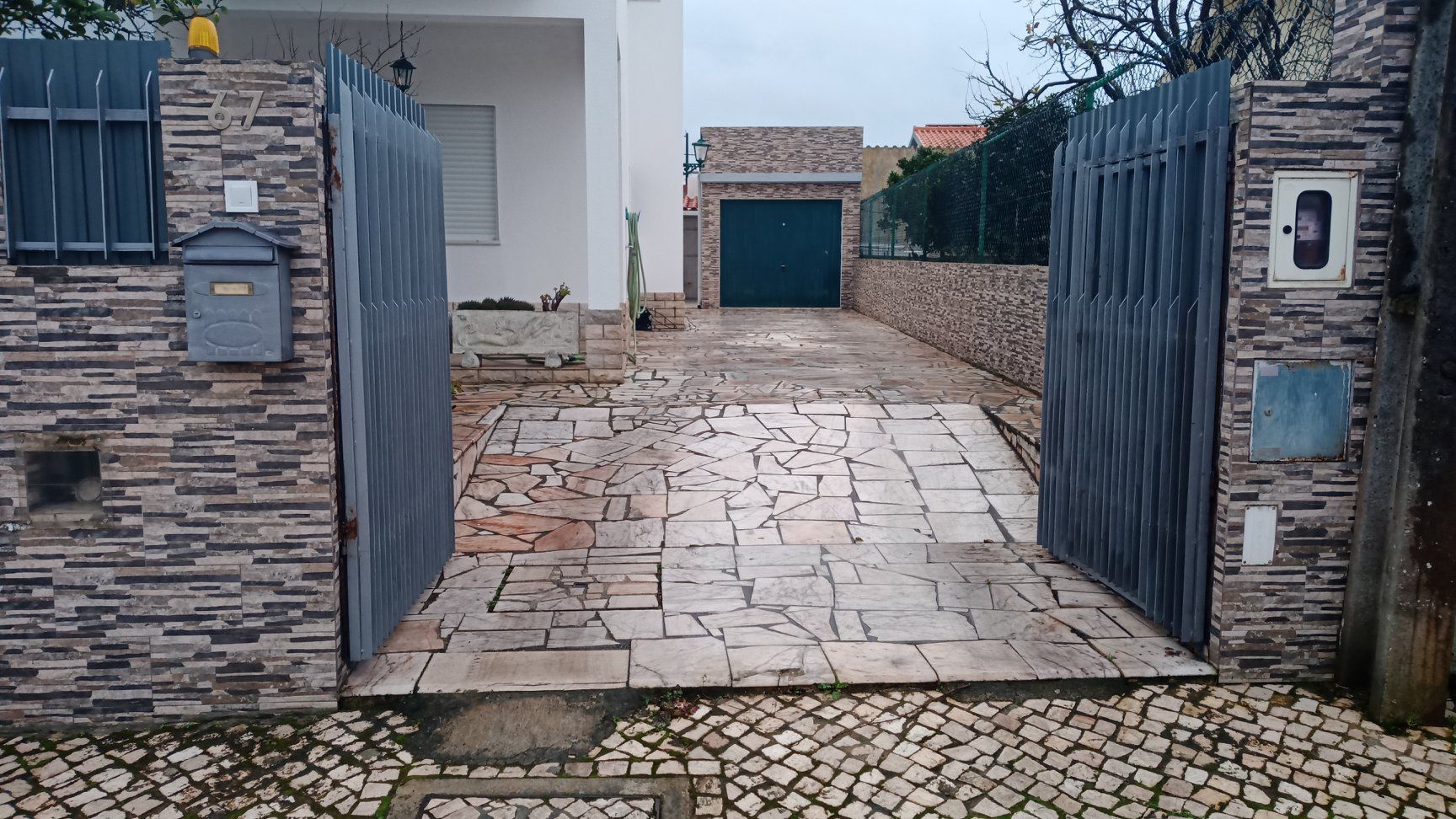 Portão de metal cinza aberto que dá acesso a uma entrada de automóveis, com a casa ao fundo.