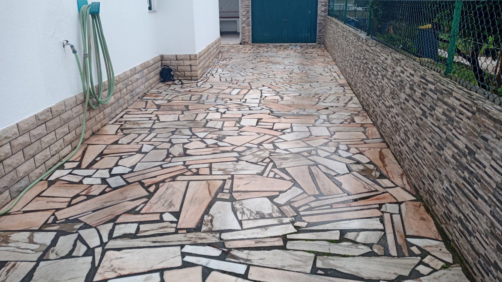 Caminho pavimentado com lajes de pedra irregulares, que leva a uma porta verde. Paredes de tijolos em ambos os lados.