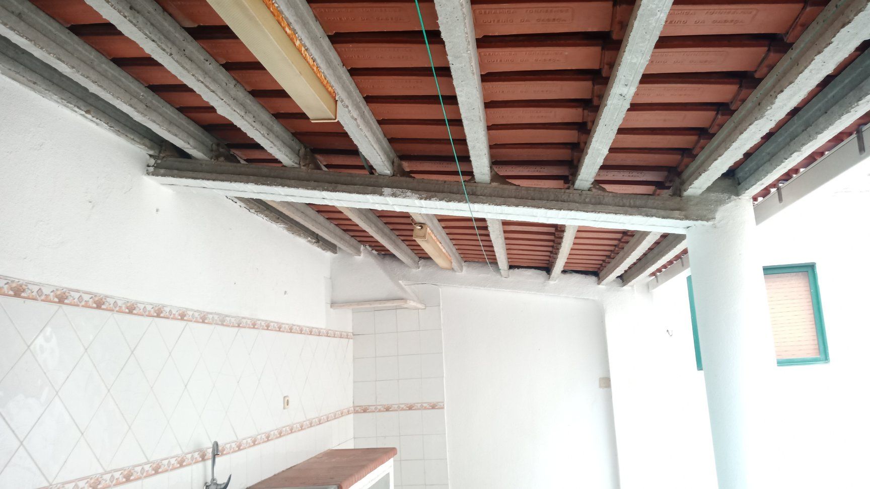 Vista de um teto com vigas de concreto e azulejos vermelhos, parte de um espaço externo com paredes brancas e uma área com pia.