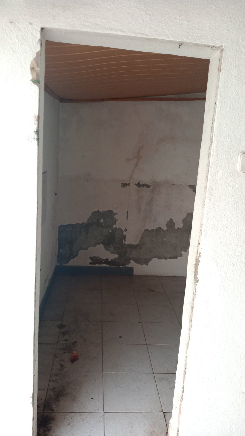 Porta que dá acesso a um pequeno quarto danificado, com paredes descascando e piso de azulejos.
