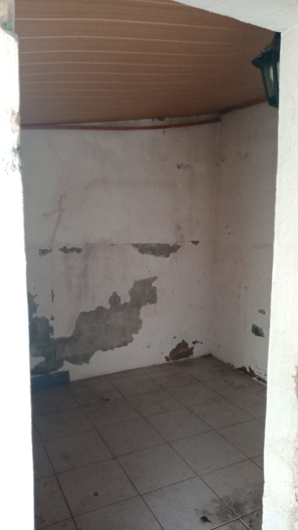 Quarto pequeno e vazio, com paredes brancas, tinta descascando e piso de azulejo, visto da porta. Teto marrom e luz externa visíveis.