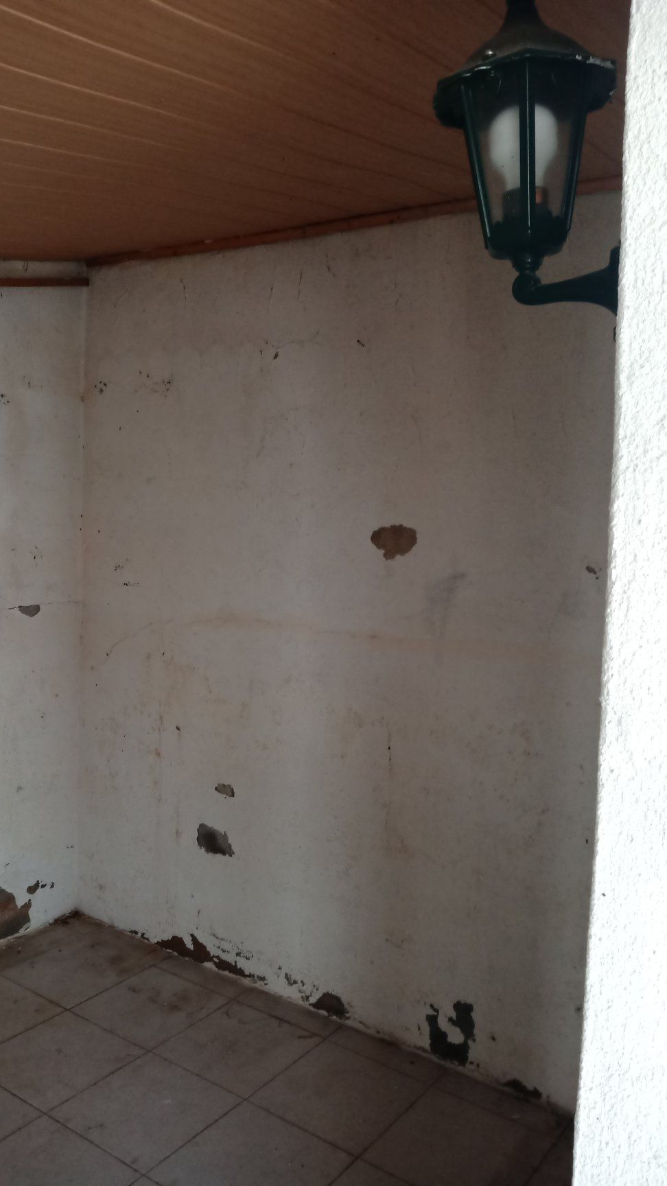 Canto de uma alcova desgastada e pintada de branco, uma lâmpada fixada na parede e piso de azulejos.