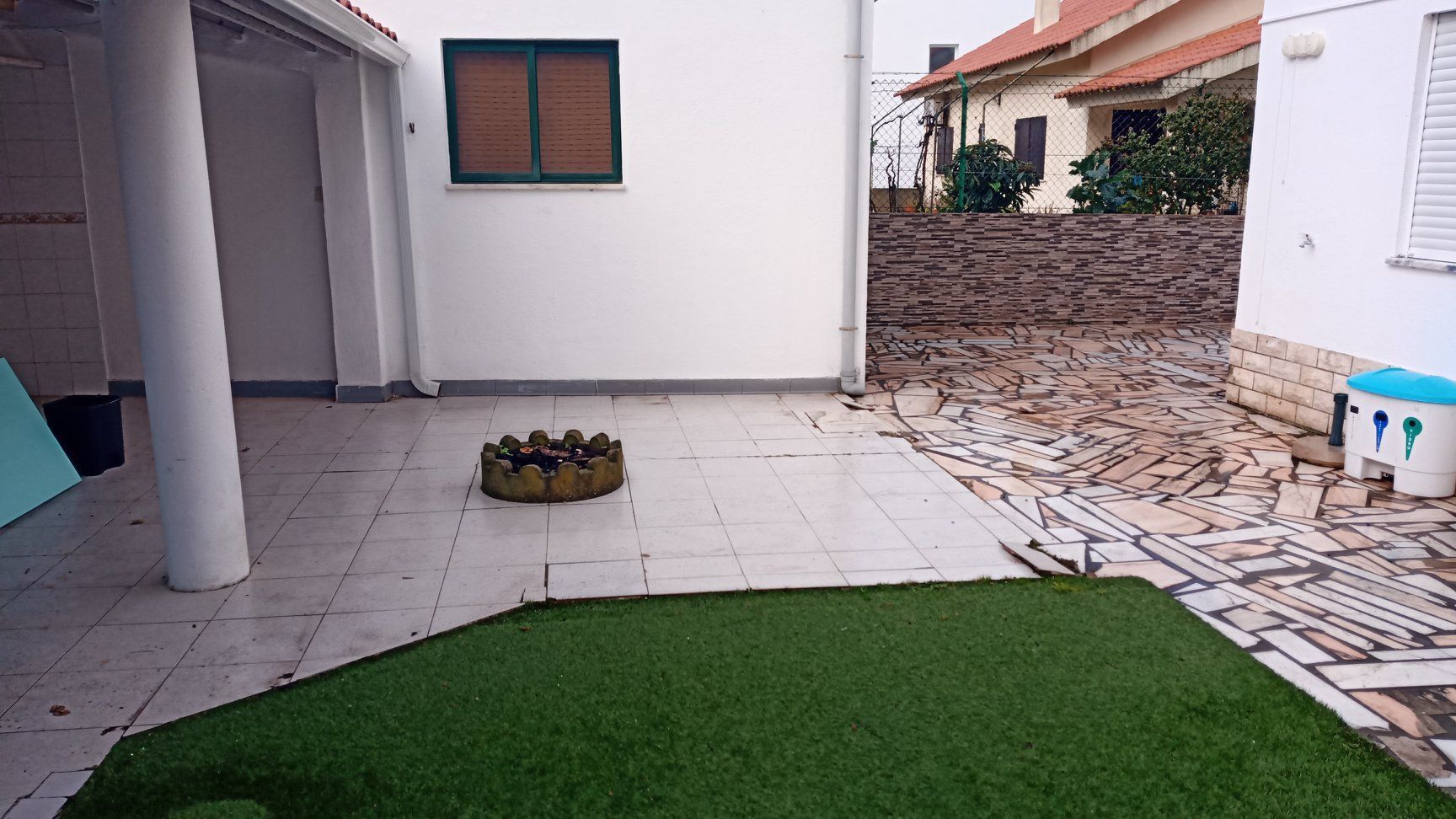 Quintal com grama artificial, lareira externa, paredes brancas e uma janela.