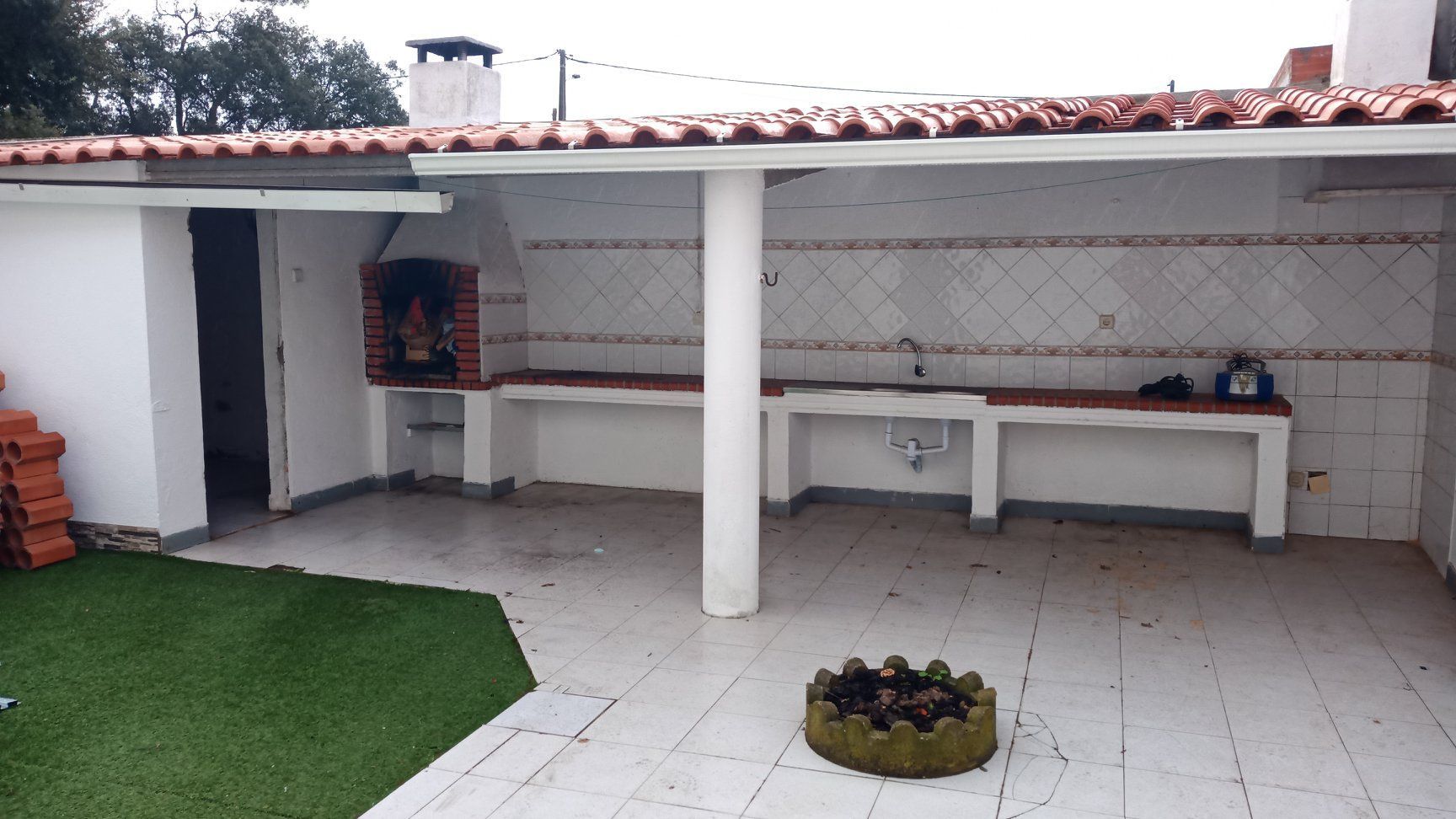 Cozinha externa com churrasqueira embutida, bancada, pia e telhado de telha vermelha. Detalhes em branco e vermelho. Grama artificial.